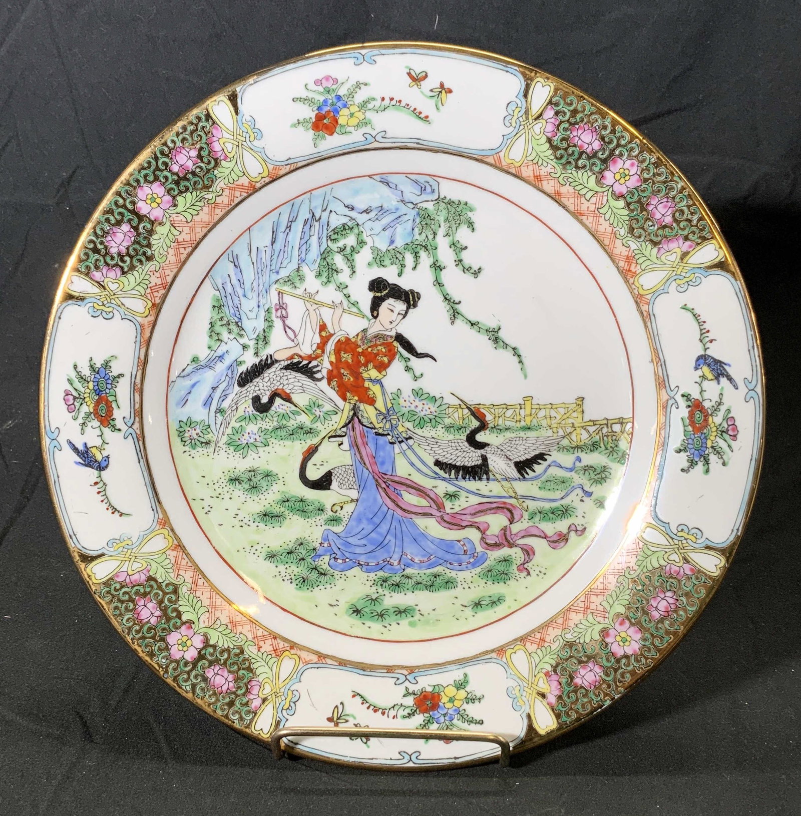 Hand Painted Chinese Figural Porcelain Art Plate: A hand painted gilt Chinese porcelain display plate. Center features female figure playing musical instrument with cranes. Border features decorative floral pattern. Hues of green, pink, and blue. Mea