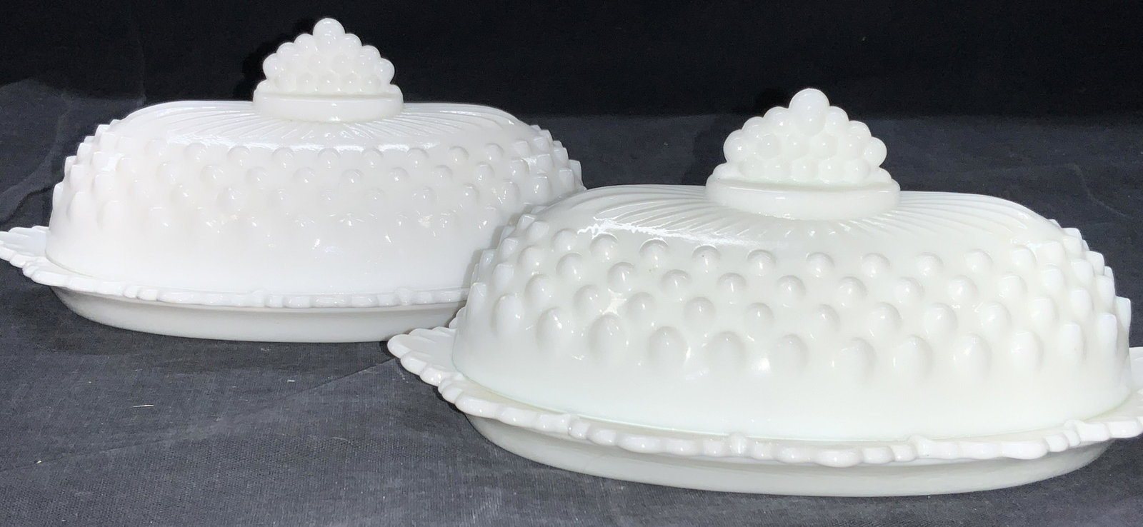 2 Vintage Fenton Hobnail Milk Glass Butter Dishes: Pair of Fenton Hobnail milk glass butter dishes. Dishes to sets have rims that resemble leaves and covers are ribbed with sunburst grooves on top, with decorative grape handles on topside. Approximate
