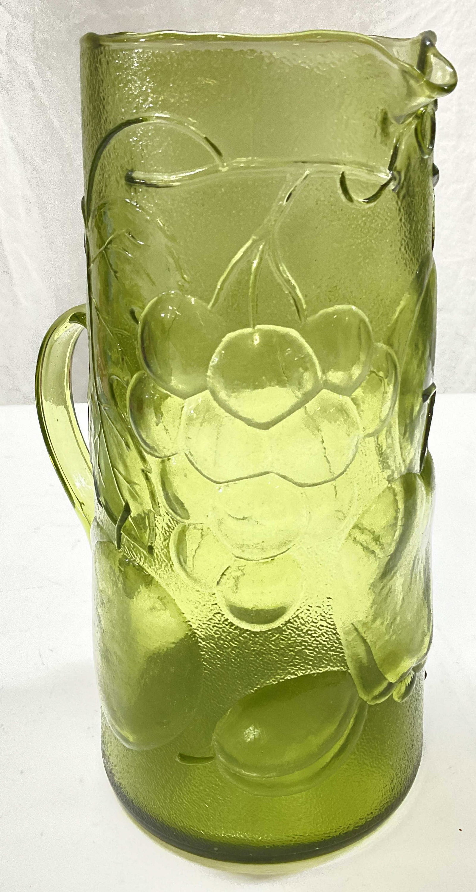 Green Glass Pitcher W Fruit Detail: Green Glass Pitcher W Fruit Detail. Measures approx 9.25 x 6 inches. Bottom reads made in Italy. Green textured glass with grape, apple, cherry & other fruit Detail to exterior. Surface wear to piece 