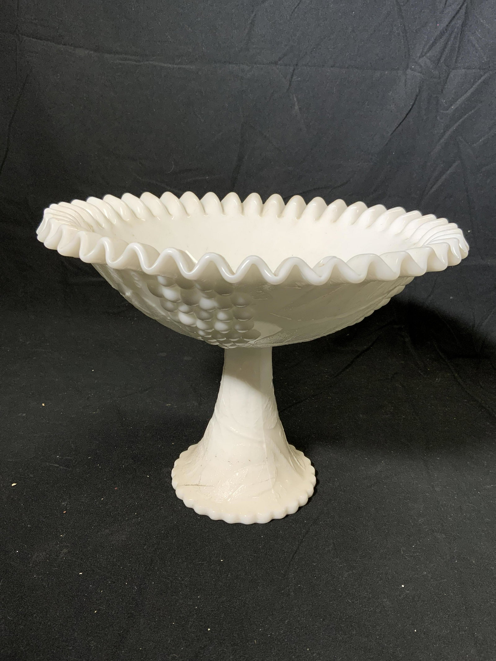 Vintage Embossed Milk Glass Grape Motif Compote: A vintage embossed milk glass compote with grape motif and furled rim. Measures approx. 10.5 inches diameter and 8 inches tall. Surface wear consistent with age and exposure. Milk glass, tureen, glass