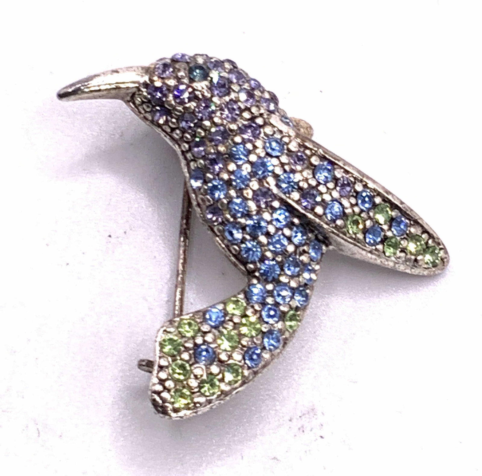 MONET Vintage Bejeweled Hummingbird Brooch: A silver tone metal hummingbird form bird brooch. Studded with blue, purple, and green hued faceted crystals. Measures approx 1L x 1.5W in. Underside signed MONET. Underside possesses metal pin closur