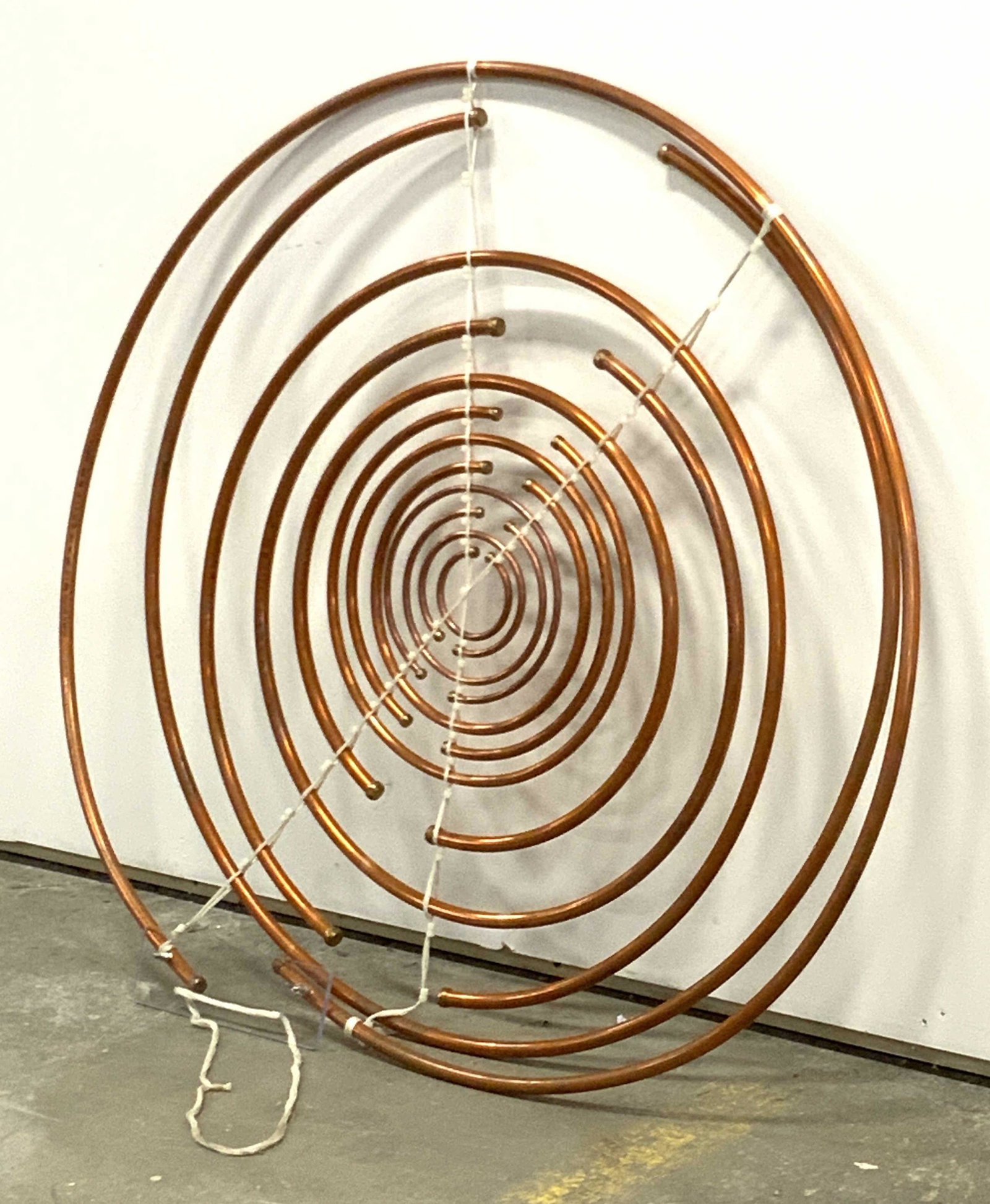 Copper Spiral Wall Sculpture: A copper spiral sculpture. Appears to be crafted from upcycled graduated copper pipes and held together by white cord. Cord is attached to a clear acrylic piece. Some pipes have lettering & numbering 