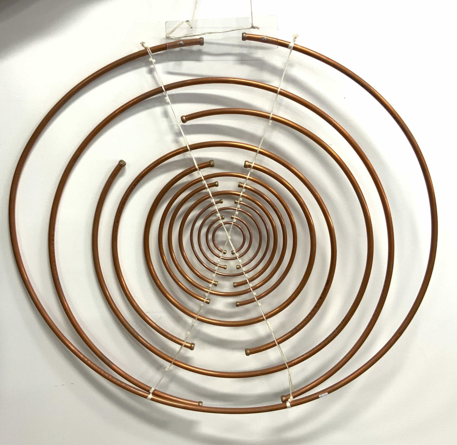 Copper Spiral Wall Sculpture: A copper spiral sculpture. Appears to be crafted from upcycled graduated copper pipes and held together by white cord. Cord is attached to a clear acrylic piece. Some pipes have lettering & numbering 
