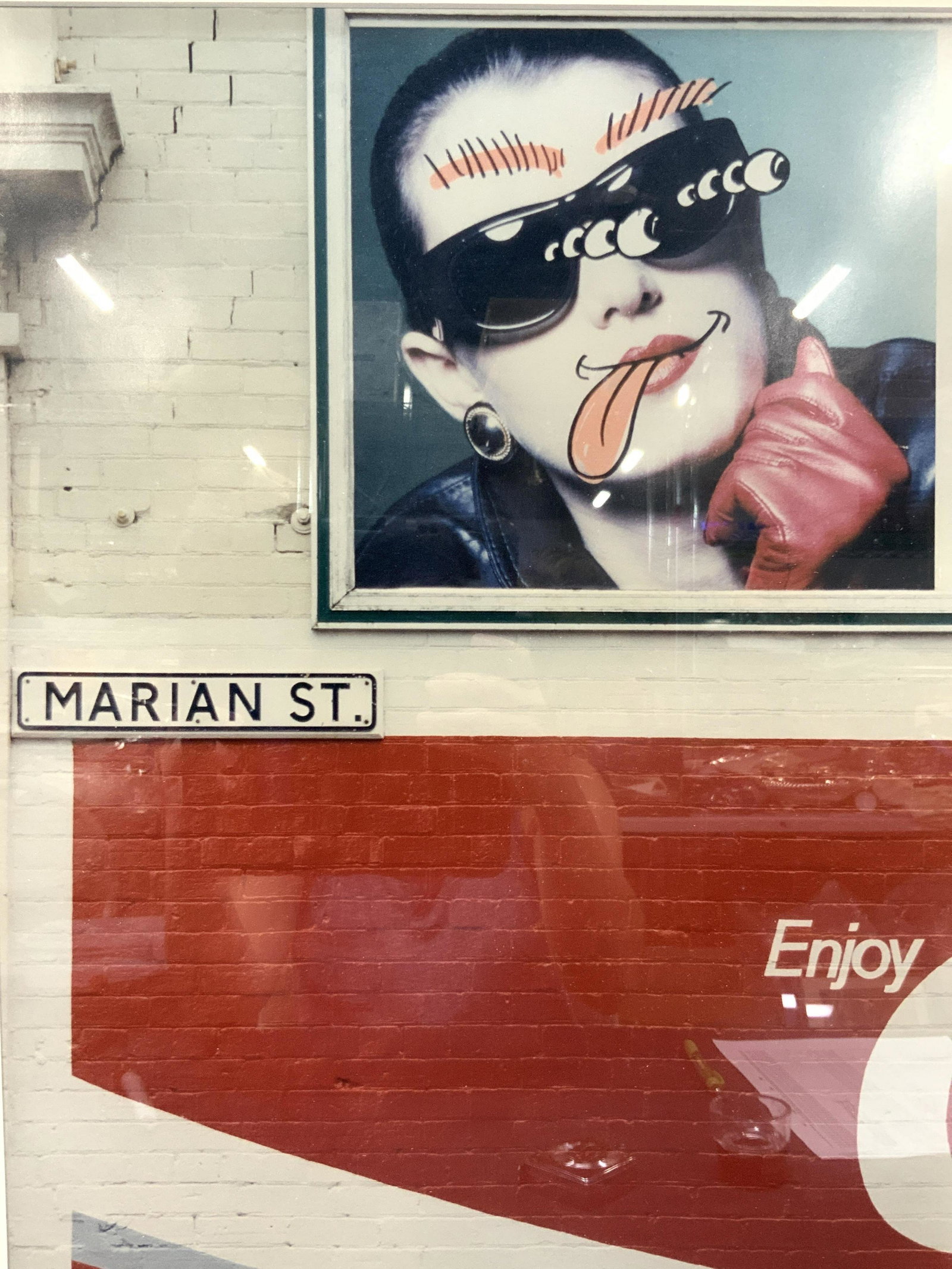 Framed Pop Art Marian St Photograph: Framed photograph depicting a brink wall painted red and white with a pop art bill board of a woman with drawn on crazy eyes and tongue. Street sign on wall reads Marian St. Approximately 2 ft x 20 in
