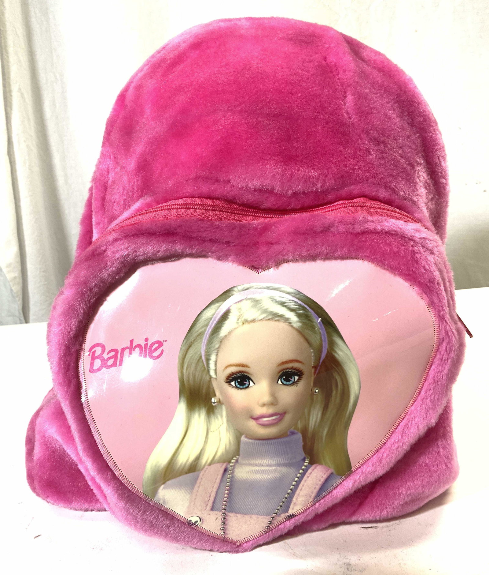 BARBIE Plush Backpack In Pink: Backpack reads BARBIE with image of Barbie Doll inside heart shape. Piece is pink toned with plush exterior. Piece has 2 zippered pockets. Measures approx 12 inches tall 10 inches wide 8 inches deep w