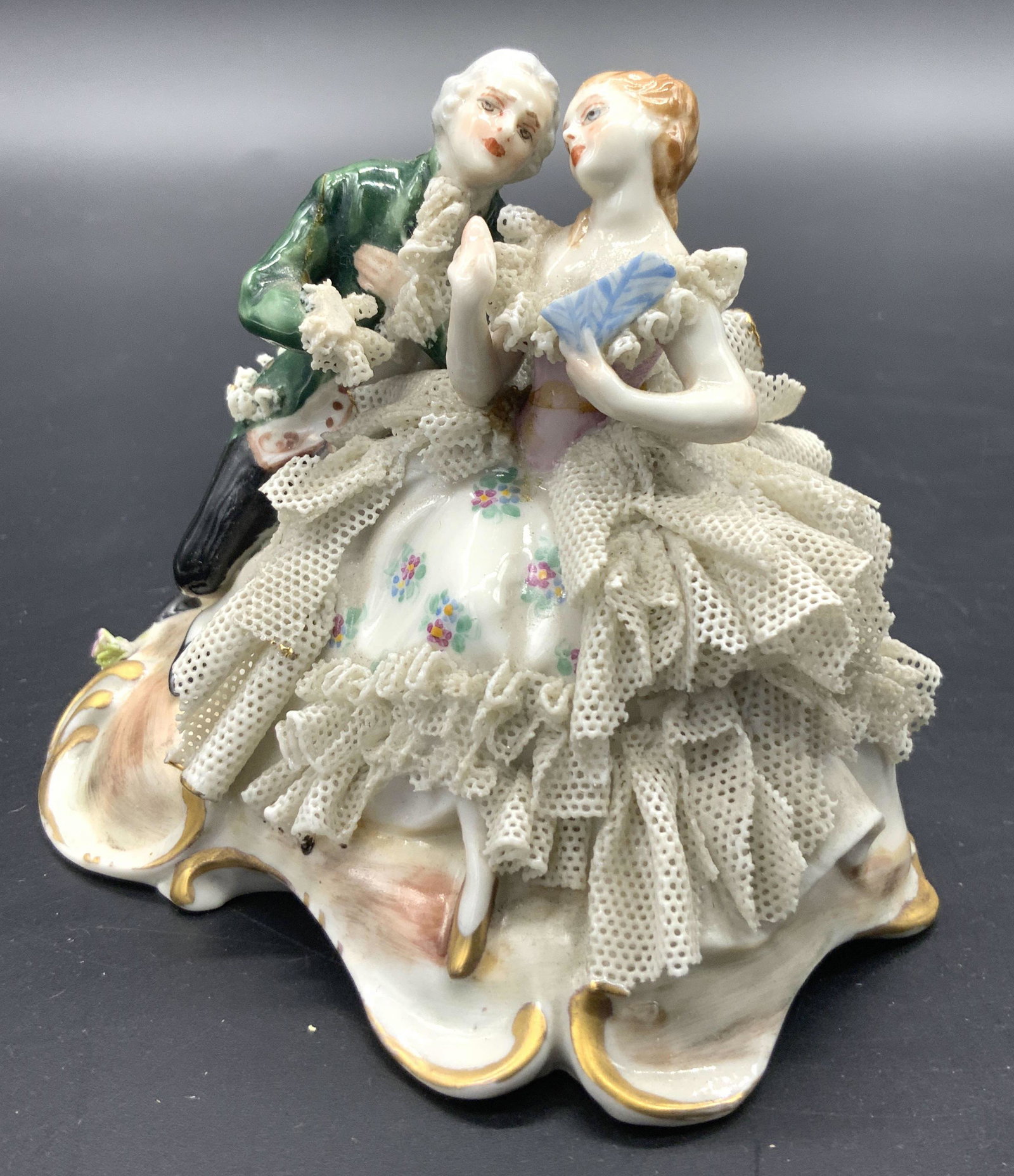 CAPODIMONTE Hand Painted Porcelain Couple Figural: A vintage hand painted porcelain figural depicting a man and a woman. Green, black, blue, and gilt hues. Florals alongside delicate porcelain work frills. Underside marked with the CAPODIMONTE tradema