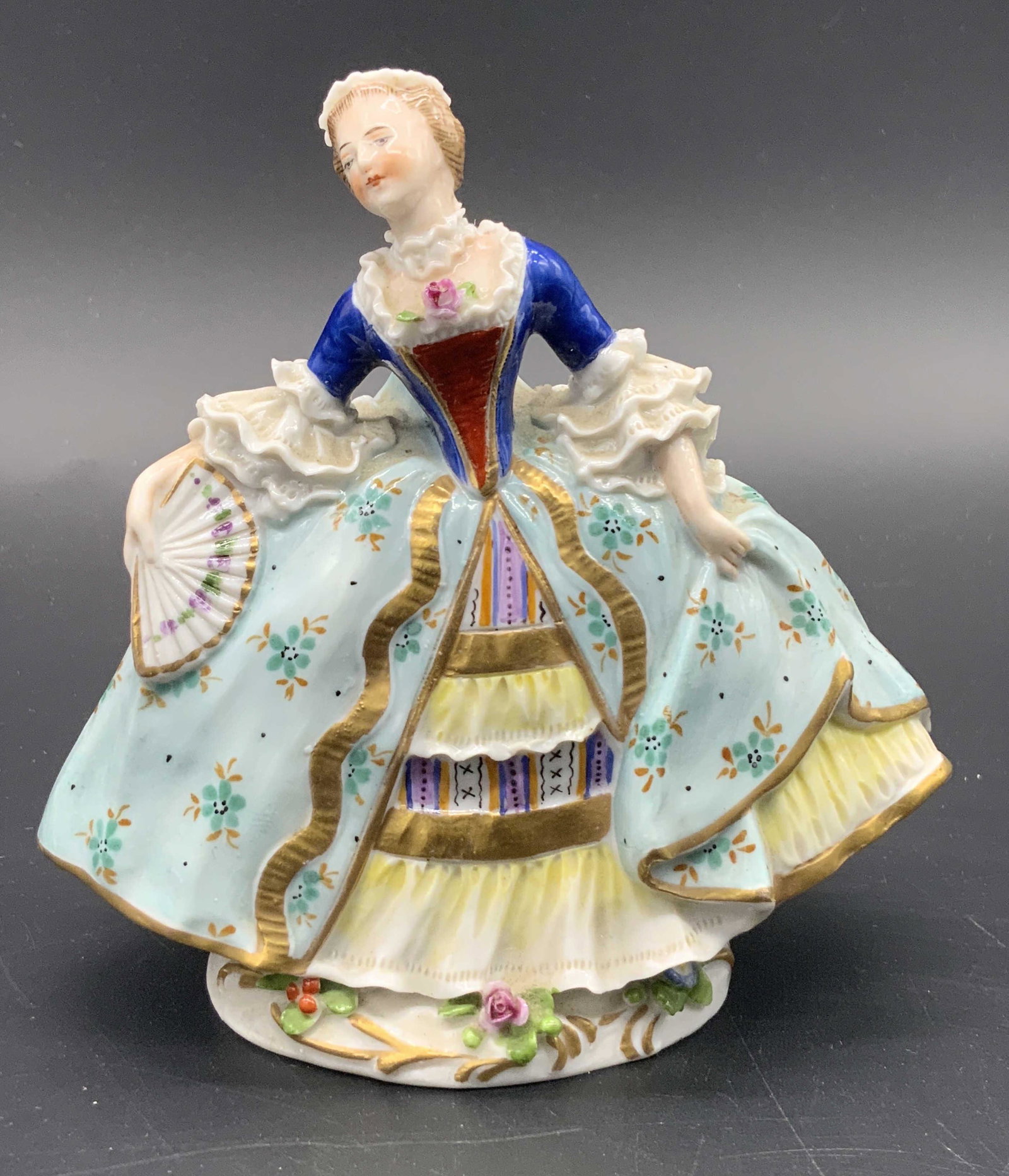 CAPODIMONTE Vintage Porcelain Female Figural: A vintage hand painted porcelain figural depicting a woman holding a fan. She wears a frilly ornate dress. Raised porcelain work throughout. Gilt, pink, blue, purple, and yellow hues. Florals alongsid