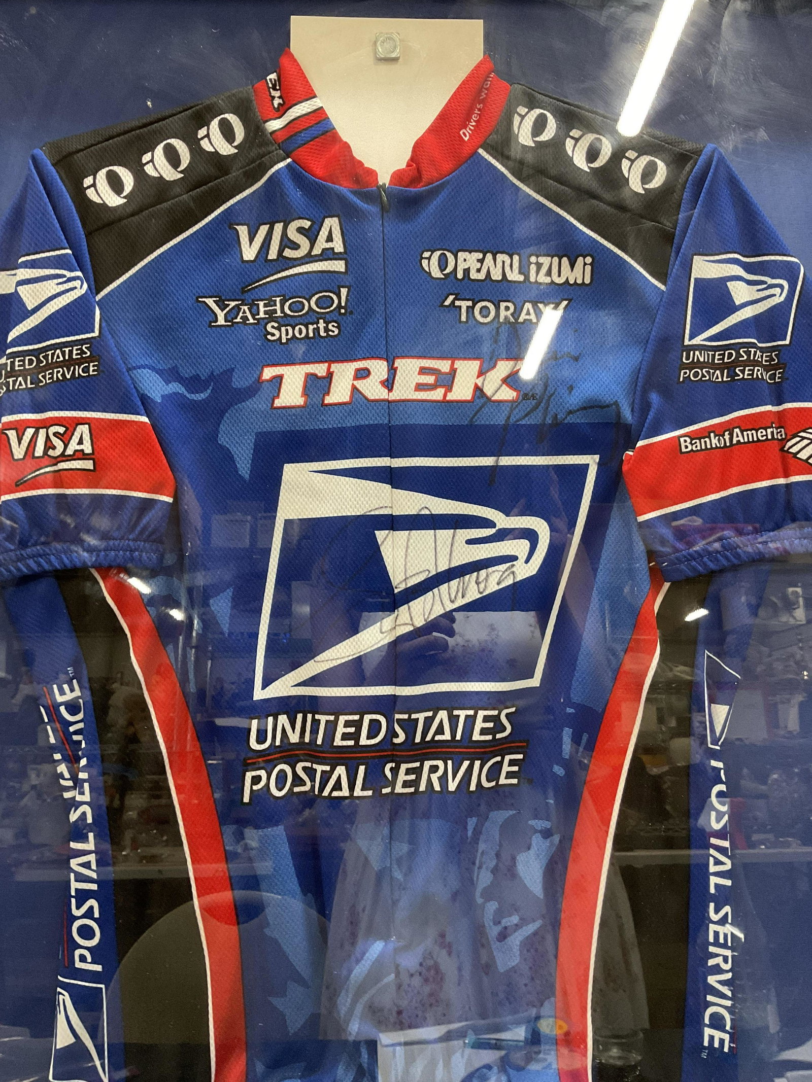 Framed Signed PEARL IZUMI Cycling Shirt: Signed PEARL IZUMI Cycling Shirt, two autographs present, neither is legible. One possibly reads Lance Armstrong. Please note The Bemefit Shop does not authenticate or fuarentee any signatures. Inscri