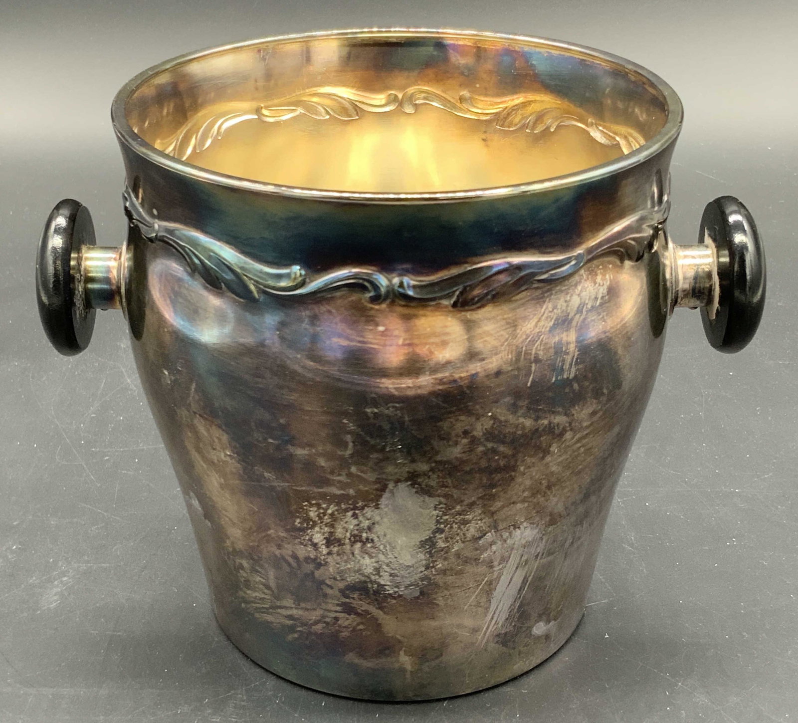 Vintage Trademarked Silver Tone Ice Bucket: A vintage silver tone metal ice bucket. Twin handles to either side, black finish to handles. Embossed scrollwork along the rim. Underside possesses a trademark. Trademarked possibly possesses letter 