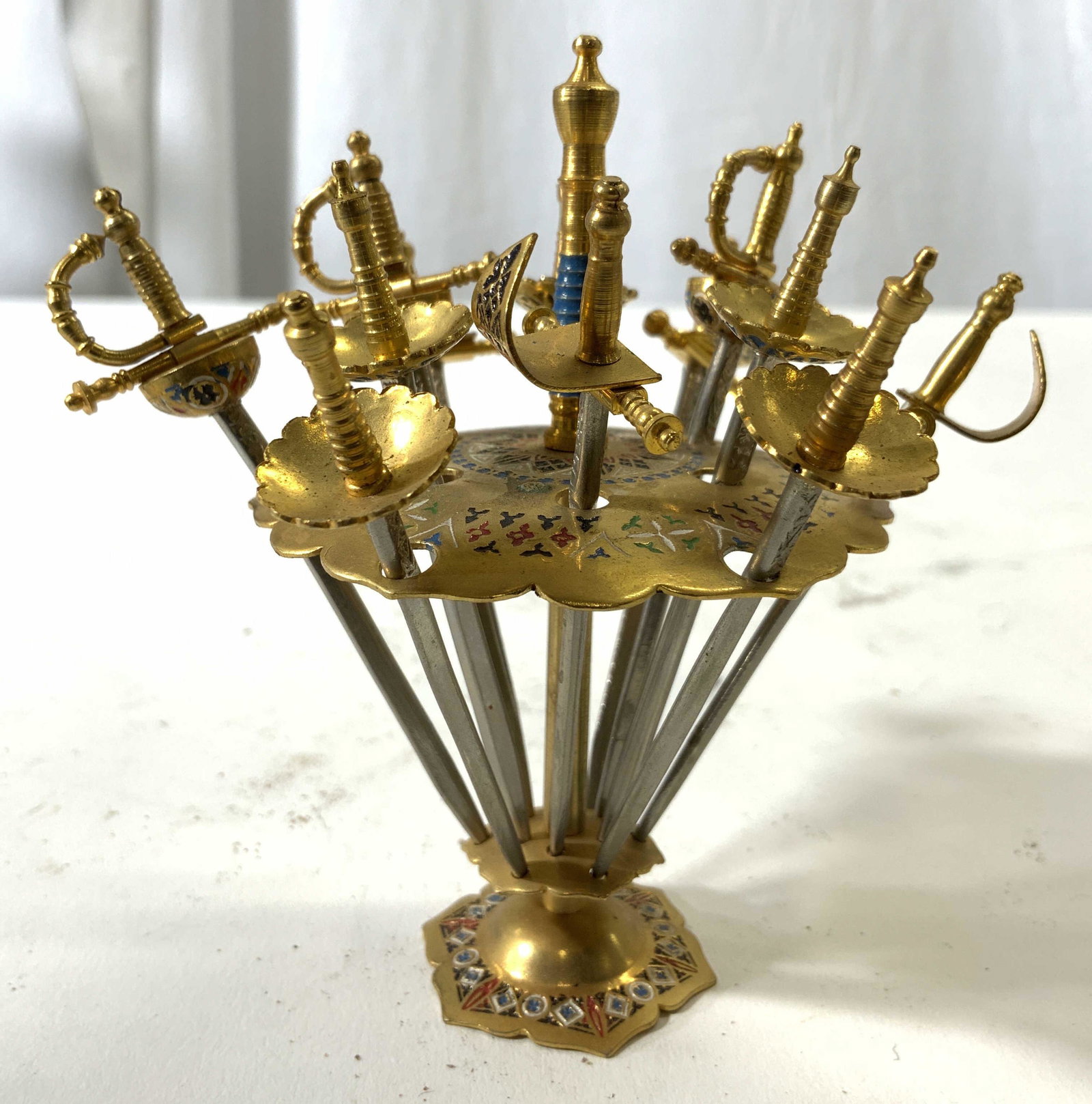 Set 13 Sword Hors D'oeuvres Cocktail Picks: Lot includes 12 sword Hors Doeuvres picks and stand. Each piece is gold toned handles, stand is gold toned with multi toned geometric detail. Stand measures approx 4 inches tall 2.5 inches wide. Cockt