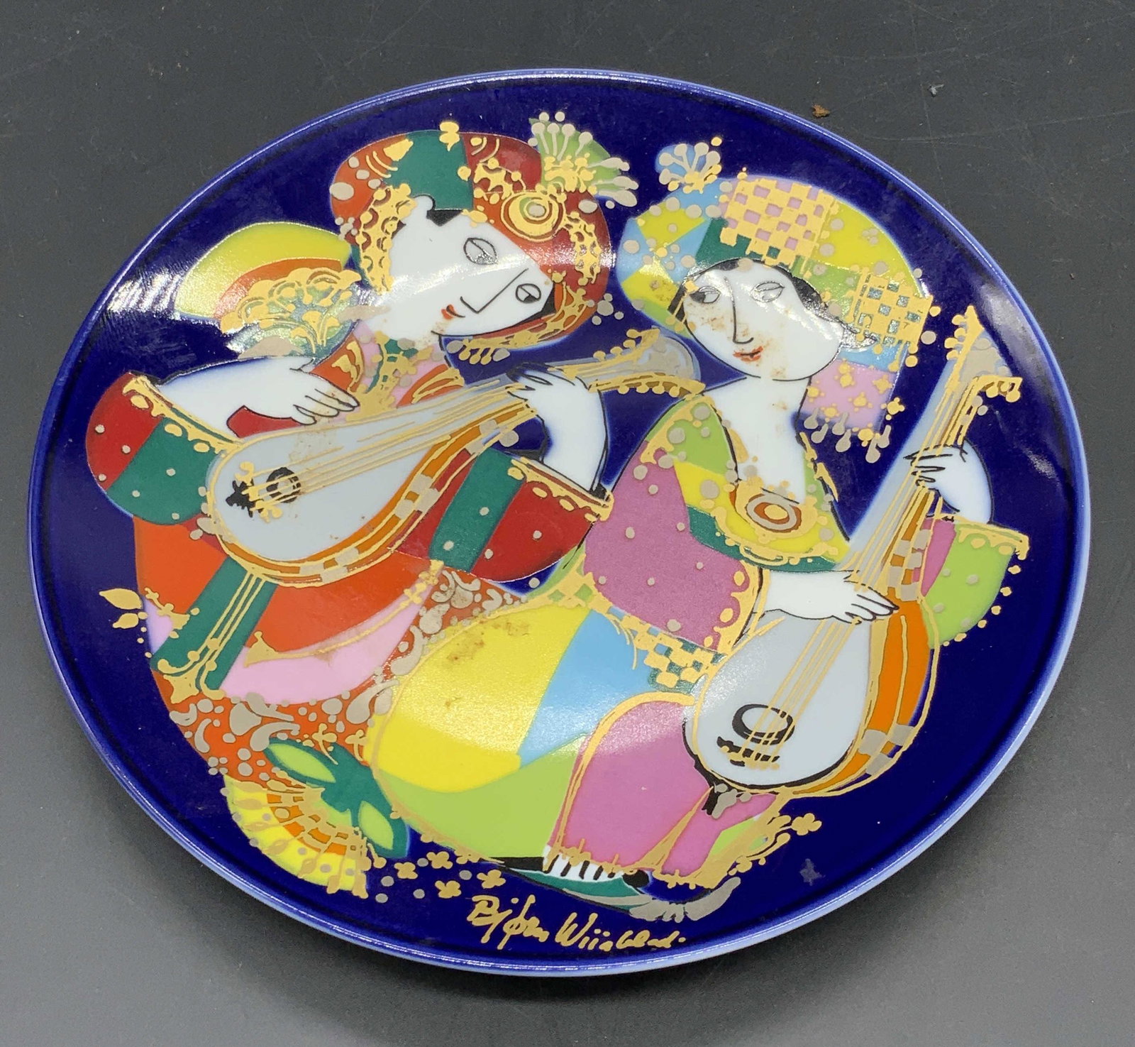 ROSENTHAL BJORN WIINBLAD Signed Porcelain Plate: A hand painted porcelain plate. Obverse so it’s two people playing mandolins. Royal blue backing with pops of bright yellow, green, red, orange, and more. Gilt accents throughout. Obverse signed BJO