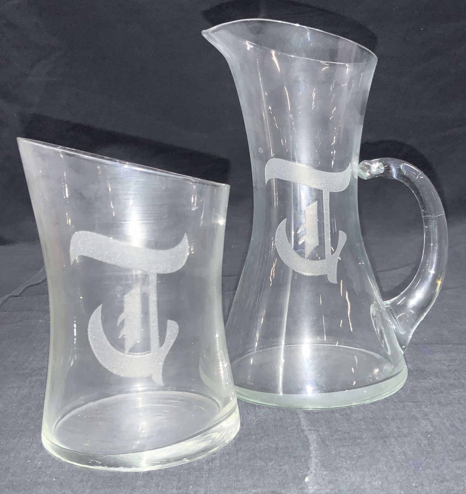 Lot 2 Etched Pitcher & Glass Set: Set of pitcher and glass with art glass shapes. Pitcher has lipped rim and side handle. Approximately 6.25 inches tall and glass is approximately 4.5 inches tall. Both etched with same symbol on exter