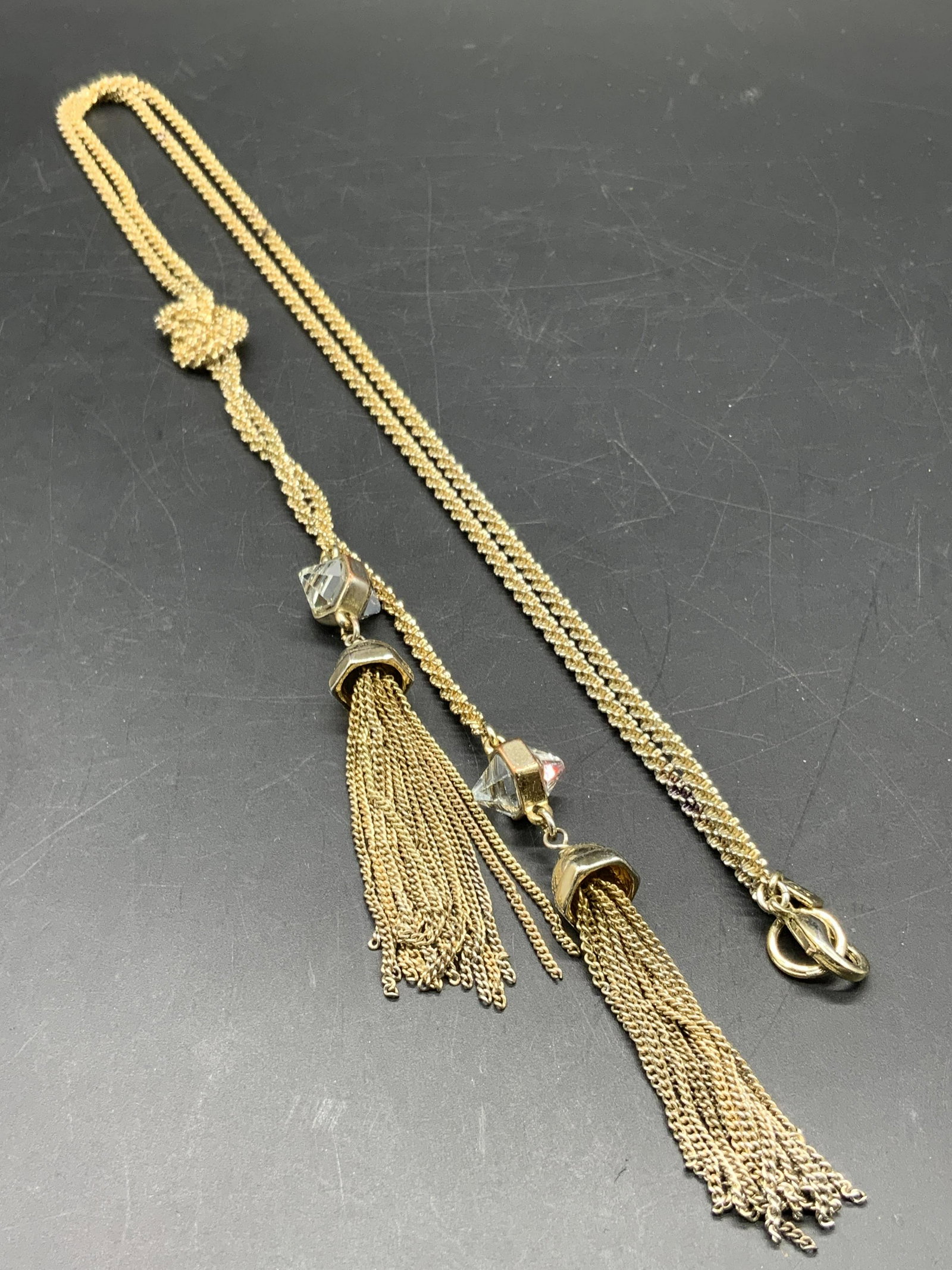 BR Knotted Tassel Pendant Lariat Necklace, Jewelry: A gold tone metal chain necklace featuring a knot to the center that two twin tassels dangle from. Faceted clear cut crystals to the tassels. Lariat style. Necklace length approx 33 in. Drop length ap
