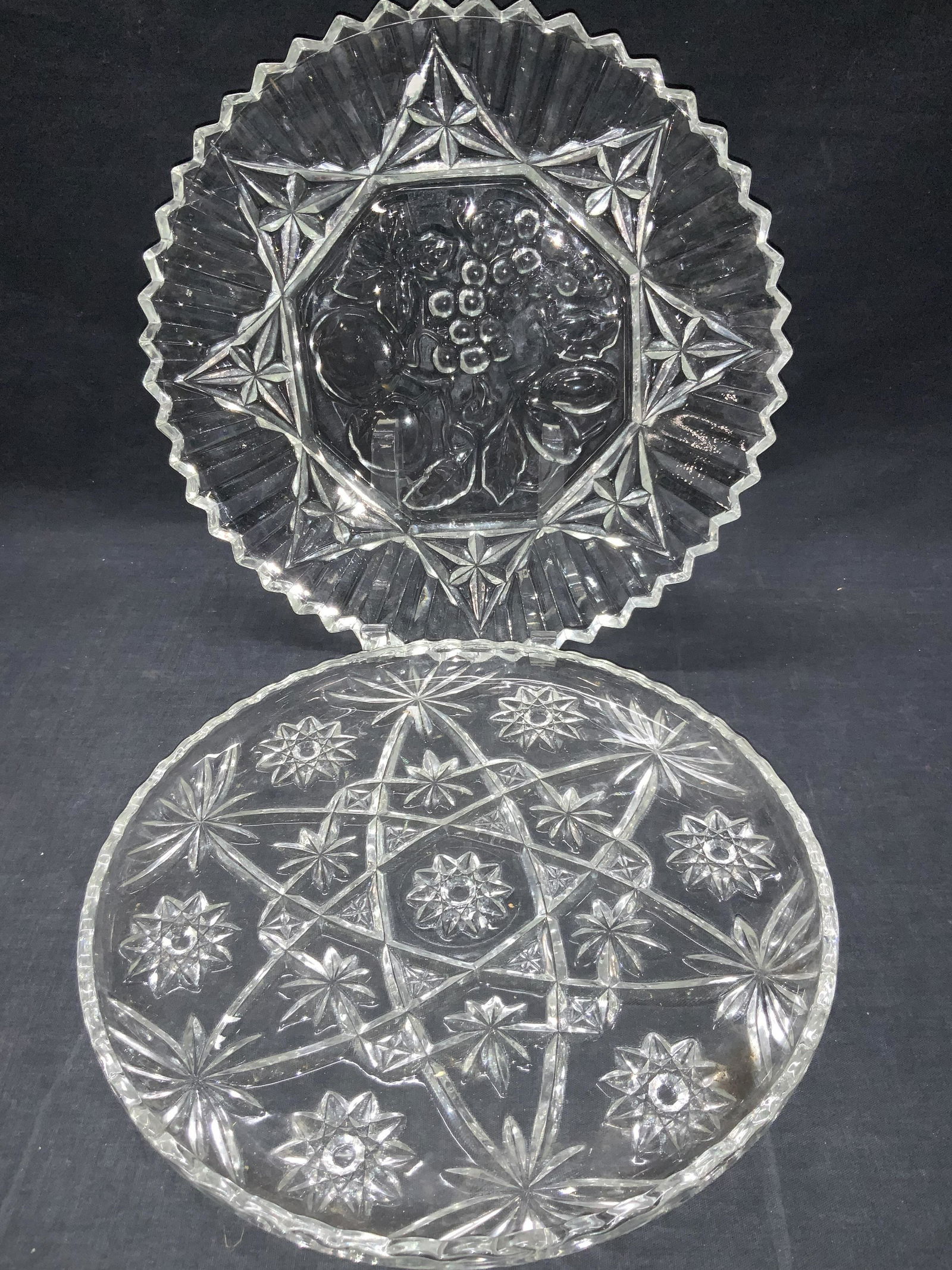 2 Clear Cut Glass Platters: 2 clear cut glass platters. 1 with embossed fruit in the center and 1 with Star bursts throughout the center of platter. Booth with saw tooth edges. Approximately 11 inch diameter. Minor wearing to pl