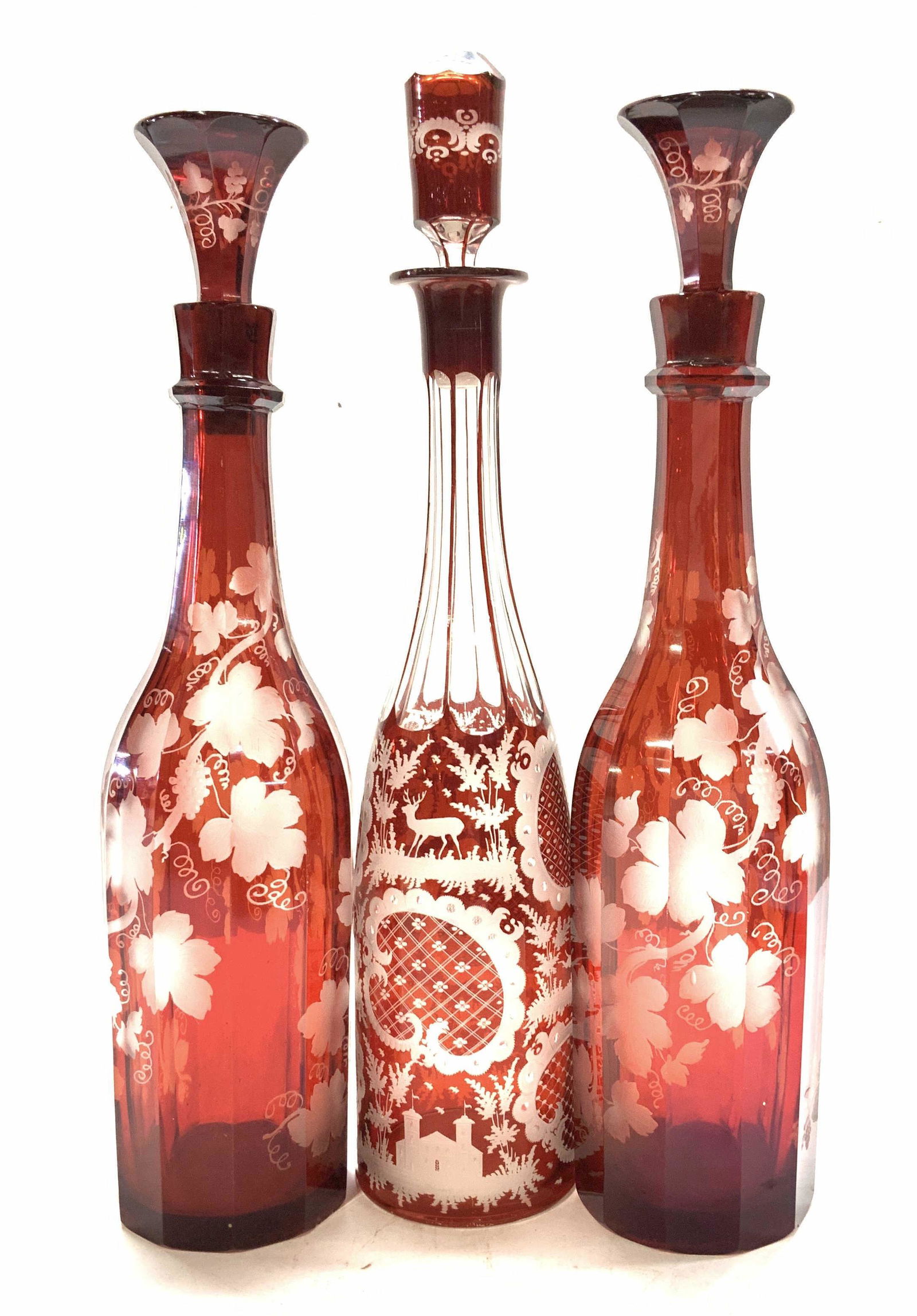Boehm Ruby Red Etched Glass Decanters, 3: 3 Bohemian Glass decanters with stoppers, etched detail. 2 have a grape vine motif with matching stoppers, the other has a classic Czech fauna with stag in field with village church. Bottles are facet