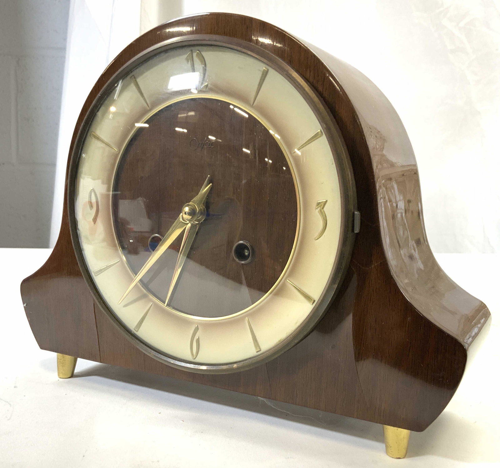 ORFAC Art Deco Wood Style Mantel Clock: Clock face reads ORFAC. Piece is brown toned and case wood style case. Clock face has gold toned hands and numbers. Piece had gold toned possibly brass feet. Piece measures approx 8 inches tall 10 inc
