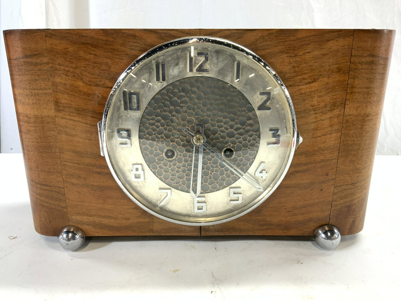 RUDOLF MOS Wood Shell Mantel Clock: Tag on interior reads HODINAR RUDOLF MOS TABORE. Mantel Clock has wooden shell. Piece has silver toned clock face. Piece has 2 silver toned metal feet in front & 2 wooden feet in back. Piece measures 