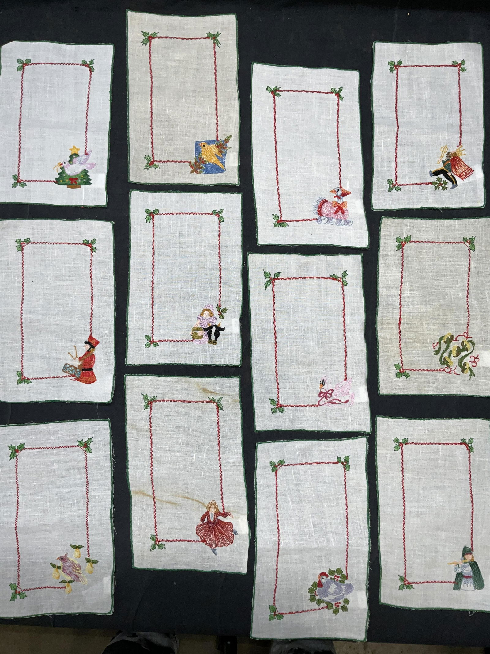 Lot 12 Embroidered Tabletop Holiday Linen Decor: Lot 12 Embroidered Tabletop Holiday Fabric Decor. Images portray the 12 days of Christmas items. Material tag reads ‘ 45% cotton 55% linen. Surface wear to some pieces, staining & minor discoloratio