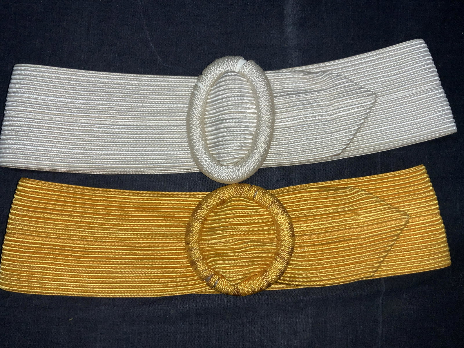 Lot 2 Vintage Colored Wide Waist Belts: 2 vintage wide waist belts. 1 white with braided oval shapes buckle and 1 yellow with braided circular shaped buckle. Both with Velcro ends. Approximately 34 inches long. Evident discoloration to whit