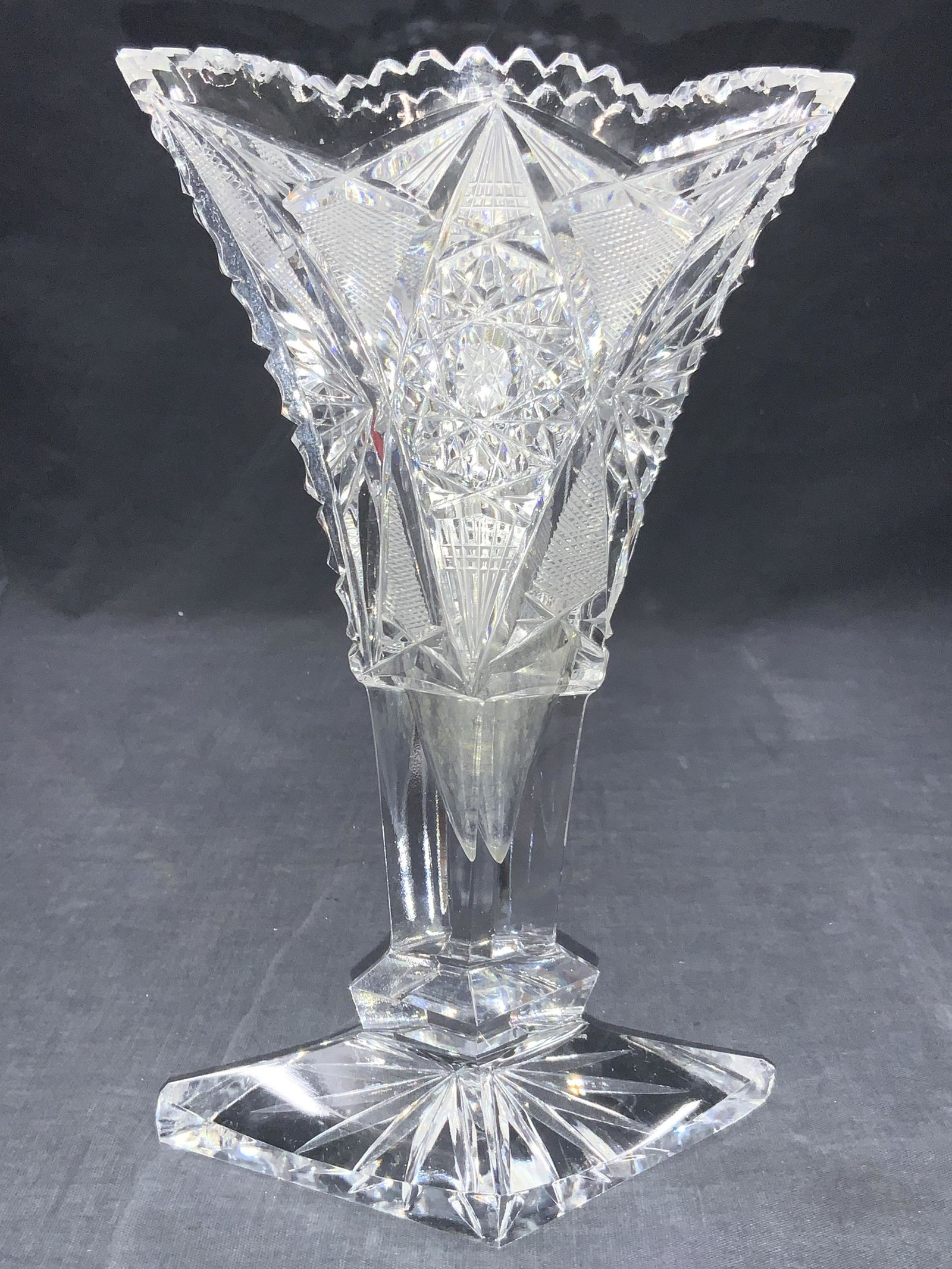 Ornate Cut Glass Footed Vase: Ornately cut glass vase with diamond shaped base and saw tooth rim. Exterior has Star Burts on centers. Approximately 8.25 inches tall and 5 inches long. Minor wearing to base corners. Ornately cut gl