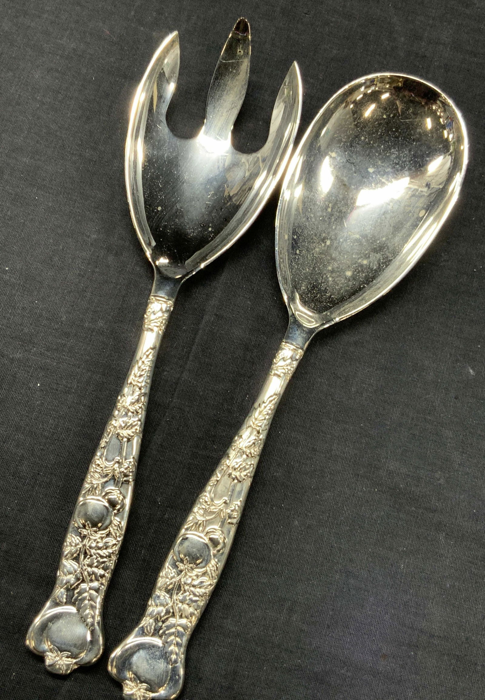 Steel Salad Serving Utensils: Pair of steel serving utensils. A spoon and 1 spork. Handles have raised designs of floral patterns. Approximately 10.25 inches long. Minor surface wear. Steel utensils, large serving utensils, utensi