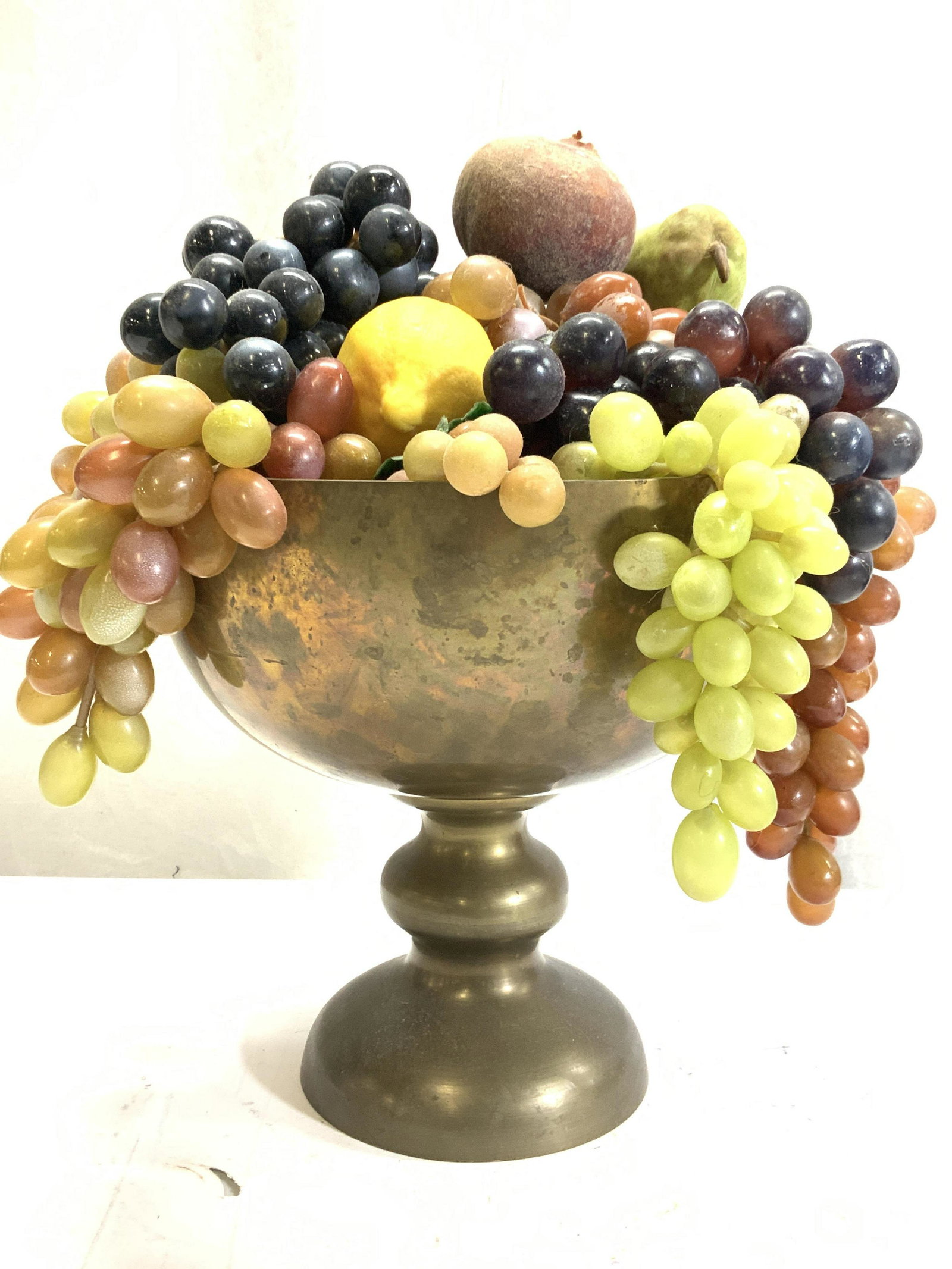 Pedestal Brass Bowl w Faux Fruit Display, India: Table Centerpiece faux fruit display in vintage brass bowl. Bowl is made in India, marked as such. Measures 9.25 x 10 in. Ht x W Green, red, and black grape bunches, a faux banana, lemon and 2 pears. 