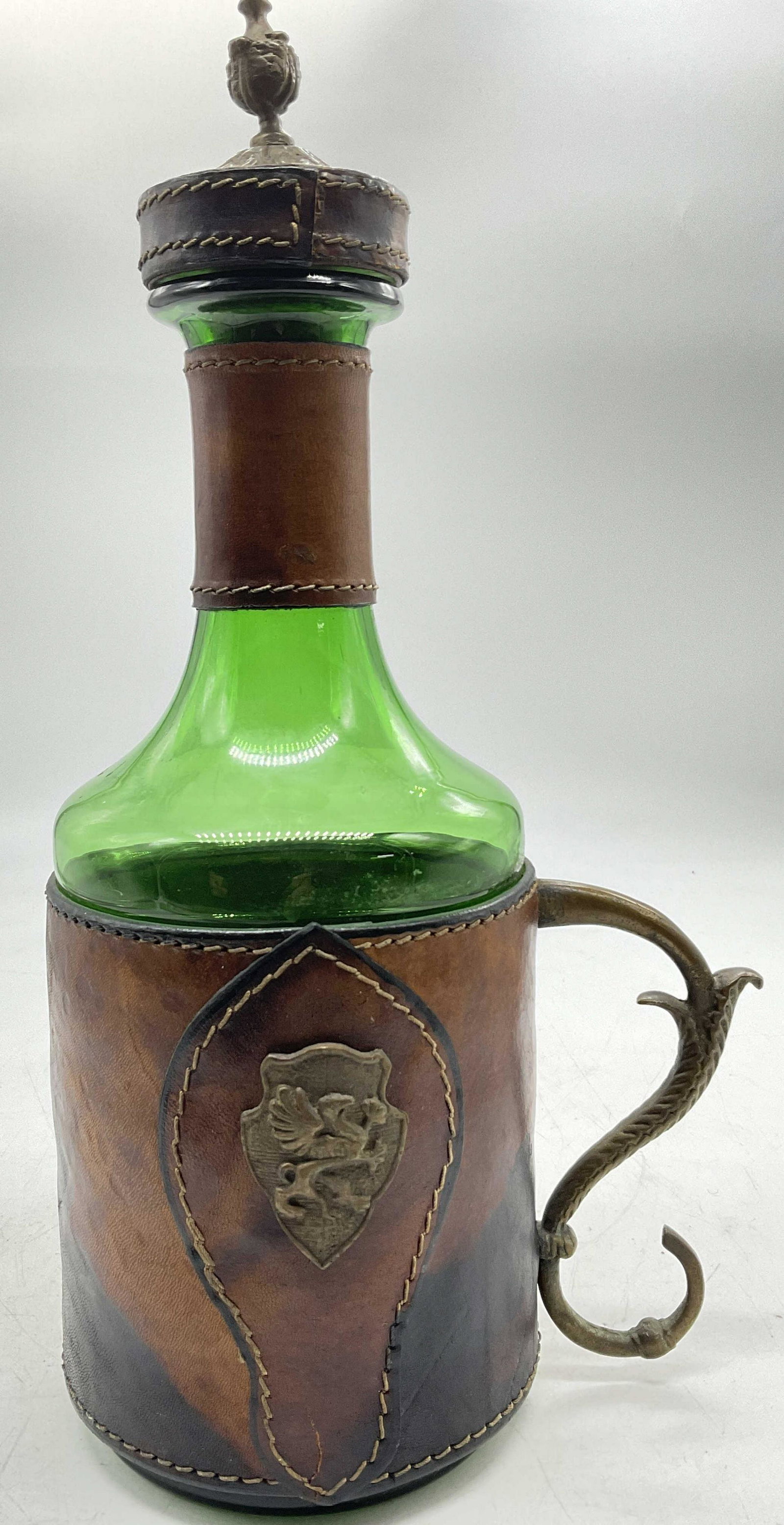 Handblown Bottle W Leather Wrapped & Brassware: Top is leather with brass ware and cork to bottom, does not close tightly to top of bottle. Body of bottle has leather wear with brass handle and brass ware with seal to front (see photos). Measures a