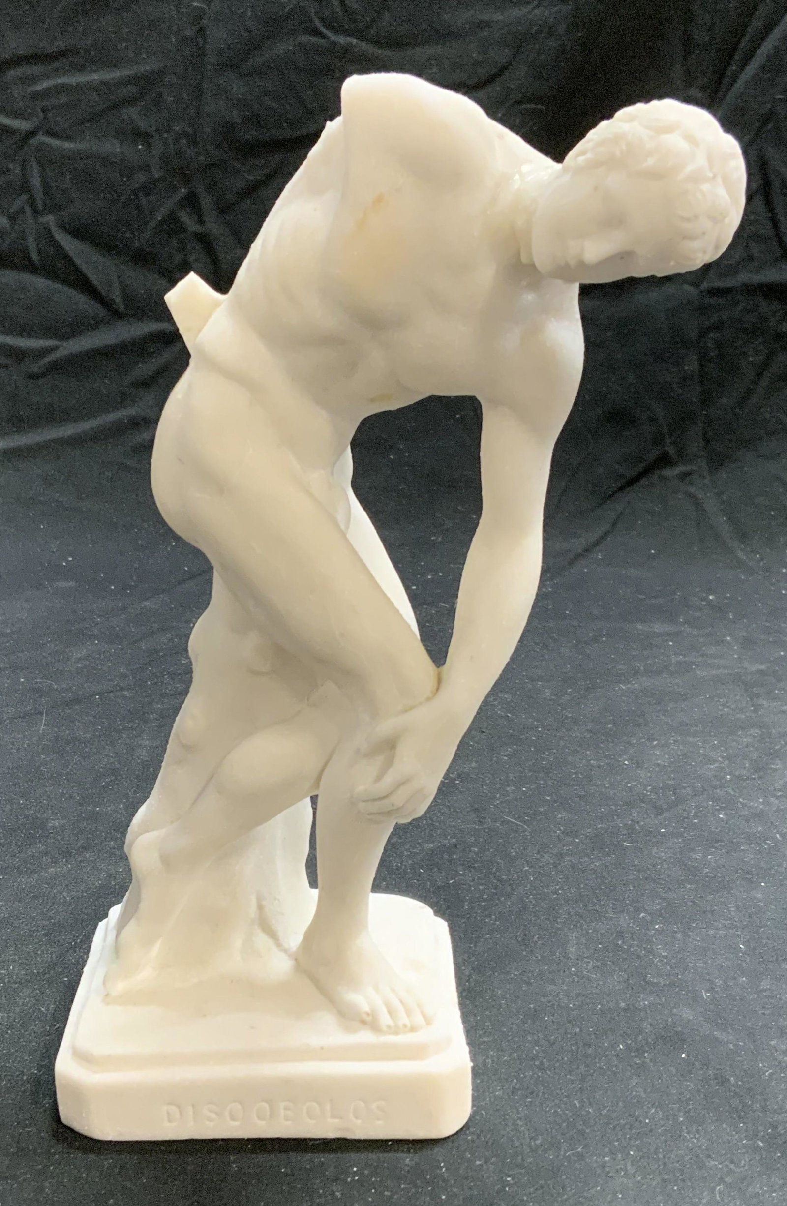 Discobolus Male Athlete Figural Table Statue: Discobolus male athlete figural table statue in white. Composite material. Piece has weight. Evident loss to arm and previous repairs observed. Measures approx. 4 x 2.75 x 7.25 inches. Discobolus, mal