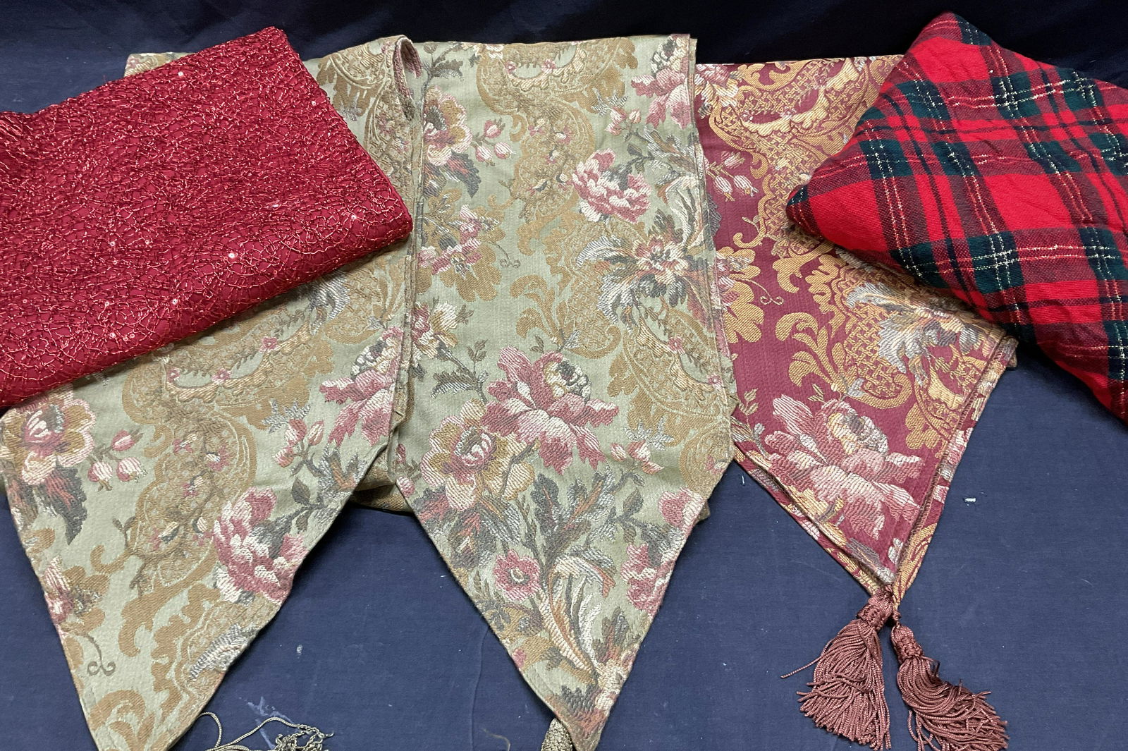 Lot 5 Tabletop Runners & Tablecloths, Holiday: 3 tabletop Floral runners with tassels to ends. Minimal wear to tassels, fraying is present. 2 holiday colored tabletop linens. Plaid and one red toned runner with sequins. Largest piece is the plaid