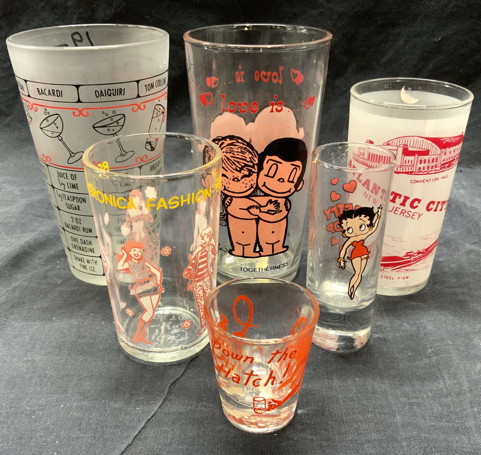 Lot 6 Assorted Vessels, Cups, Drink Ware: Lot includes 1 BETTY & VERONICA FASHION SHOW Cup, 1 ATLANTIC CITY NEW JERSEY Cup, 1 BETTY BOOP double shot glass, 1 DOWN THE HATCH shot glass, 1 Cocktail measuring cup, & 1 cup W Nude figures. Largest