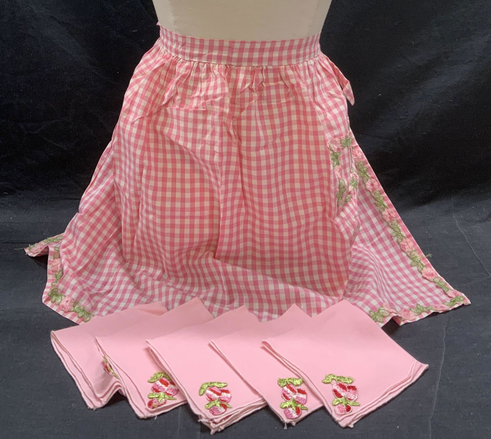 Lot 7 Pink Gingham Apron & Pink Floral Napkins: 1 pink gingham apron. Ties at waist. Waist total length measures approx. 78 inches long. Gently worn. Set of 6 pink napkins with floral applique. Measures approx. 11 x 11 inches each. Material feels l