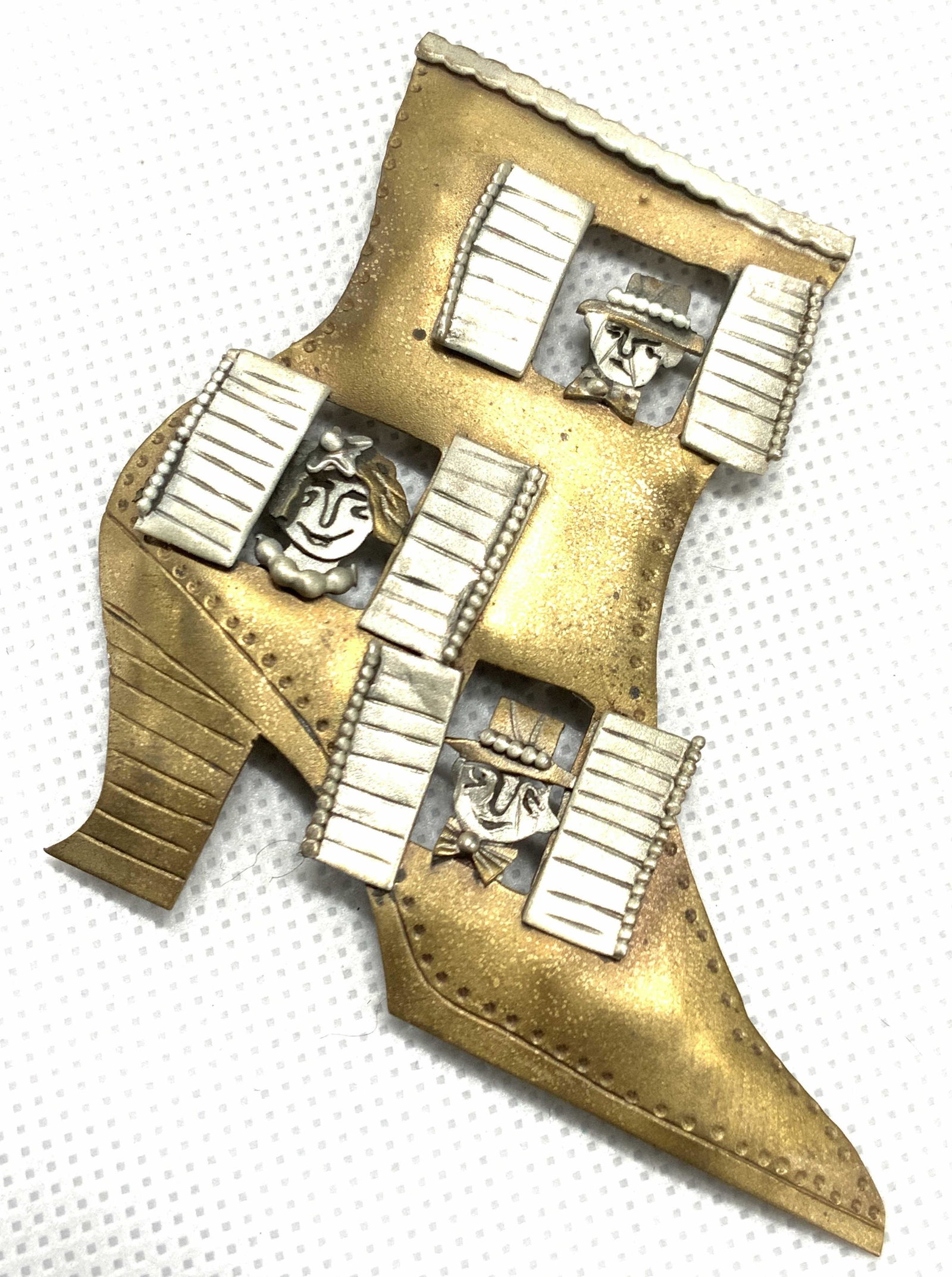 SIGNED Artisan Crafted Brass Metalwork Brooch: A hand crafted metalwork brooch. Made of brass and a white composite metal. Depicting a heeled show with three window shutters and people looking out the windows. Detail in the faces of the people. Si