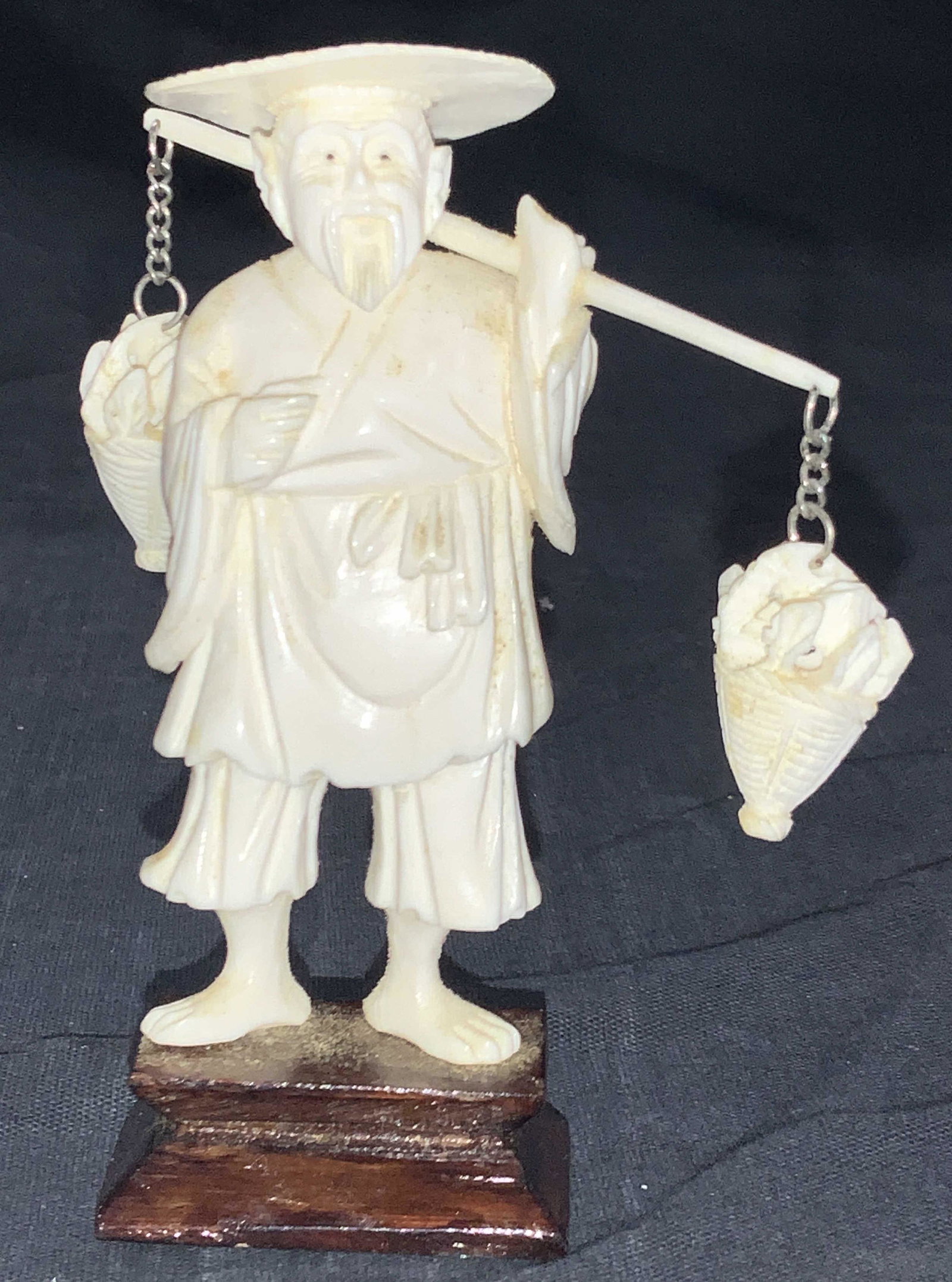 Asian Hand Carved Bone Farmer Figure: Hand carved bone figure of an Asian farmer holding a rod with hanging baskets of vegetables on either end. Farmer is wearing a straw sun hat and robes and is affixed to a wooden stand. Approximately 4