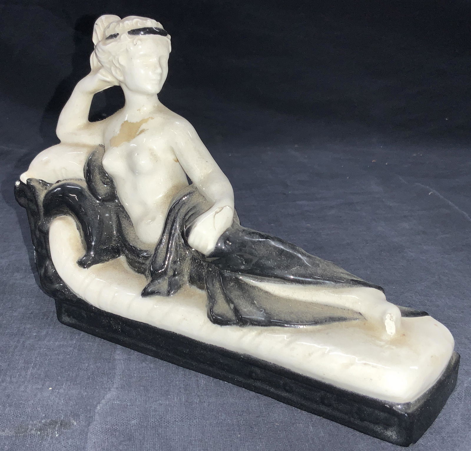 Art Deco Classical Porcelain Reclining Woman: White Porcelain reclining woman on a couch wearing a black robe. Approximately 5.5 inches tall and 8 inches long. Evident loss to figures foot and surface wearing. Art deco figure, female figure, porc