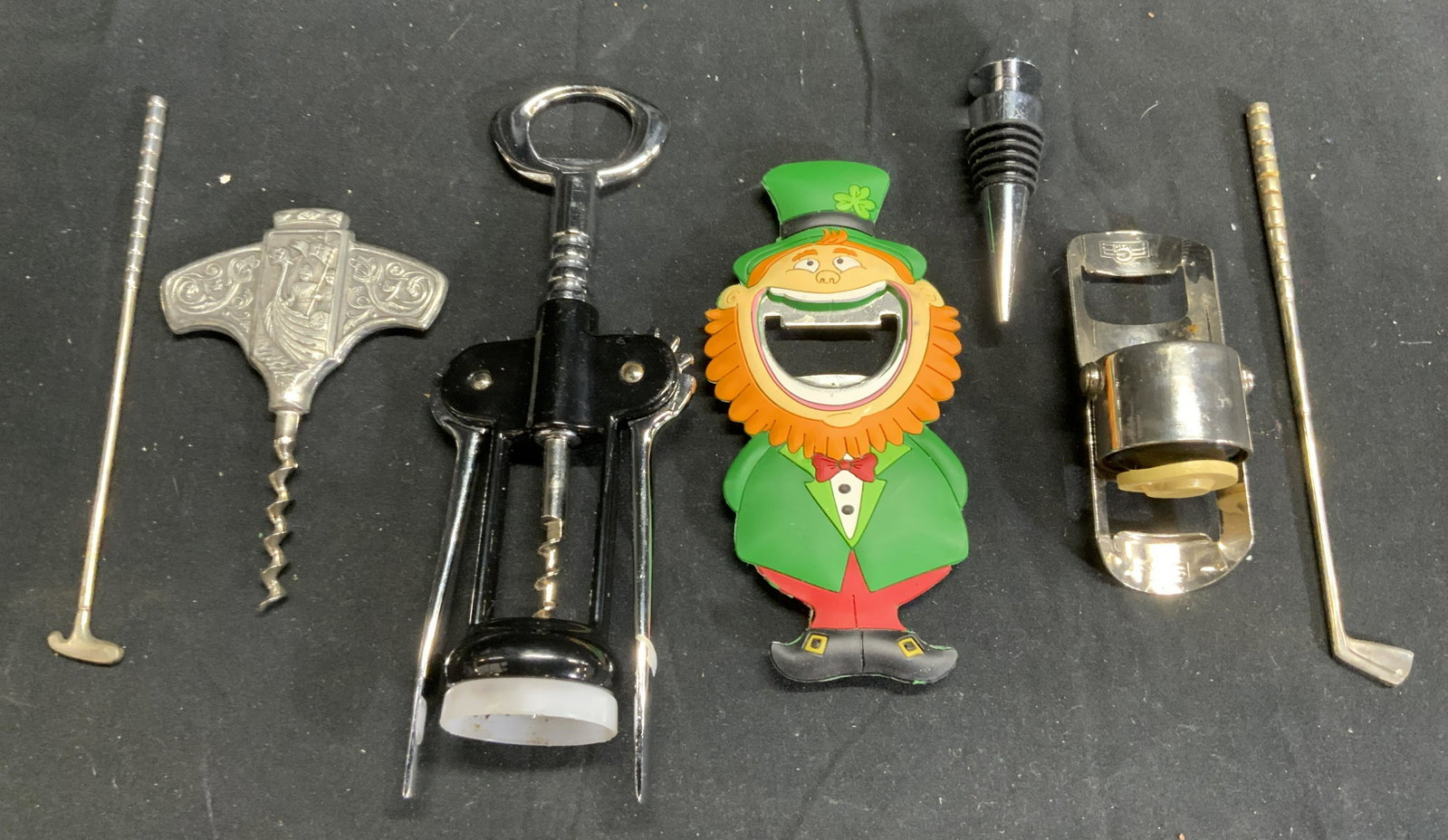 Lot 7 Assorted Barware Accessories: Lot includes 1 wine corkscrew, 1 leprechaun bottle opener, 2 golf putter figures, 1 metal wine bottle cork, 1 Pewter corkscrew & 1 FGB bottle opener. Wine corkscrew measures approx 7.25 inches tall 2.