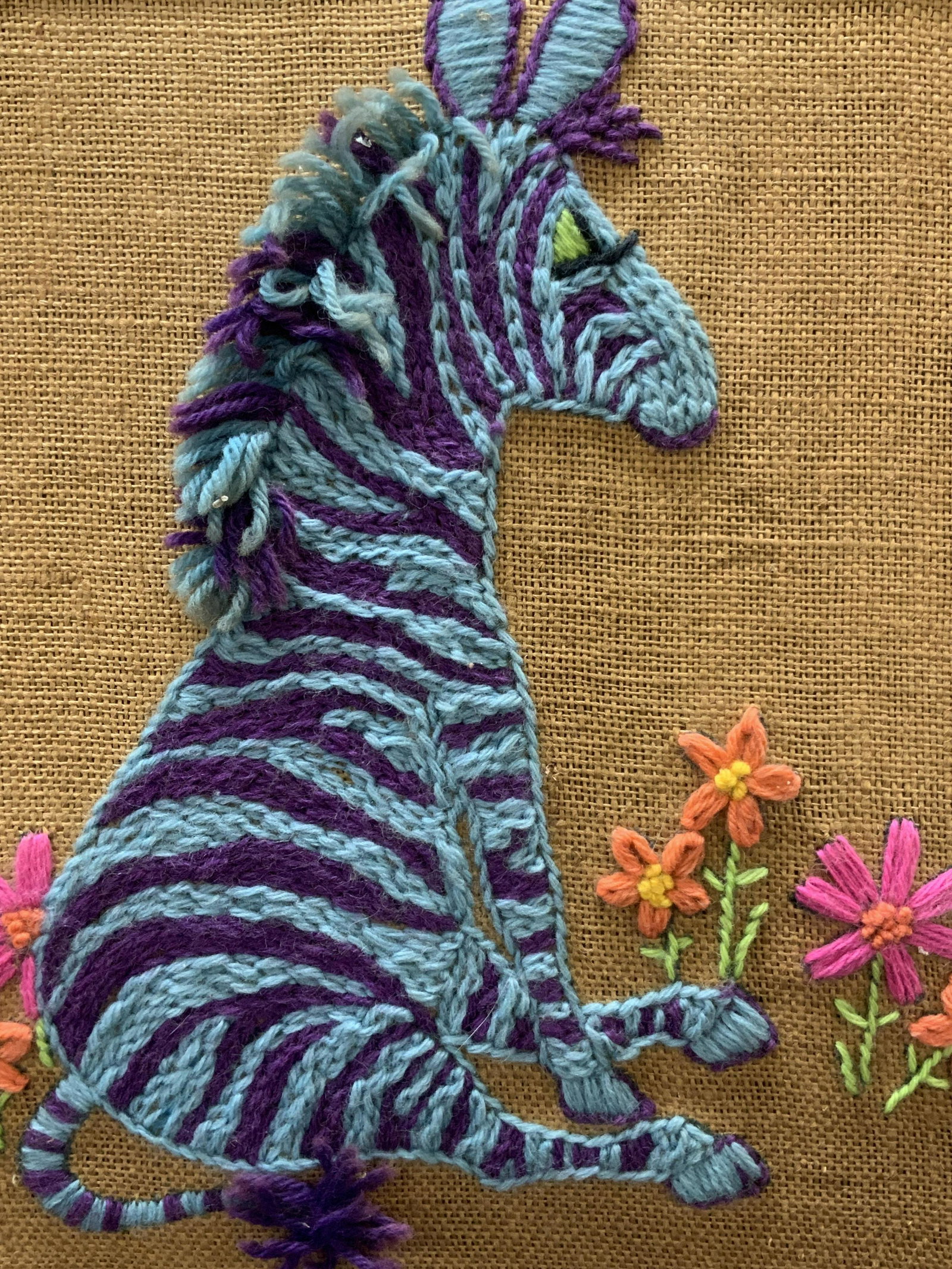 Framed Crewel Embroidery Zebra Art: Crewel Embroidery of a blue and purple striped zebra sitting amongst flowers. Framed in wooden frame Approximately 17.5 x 16.5 inches window measures 15.5 x 15 inches. Moderate wearing to frame and bl
