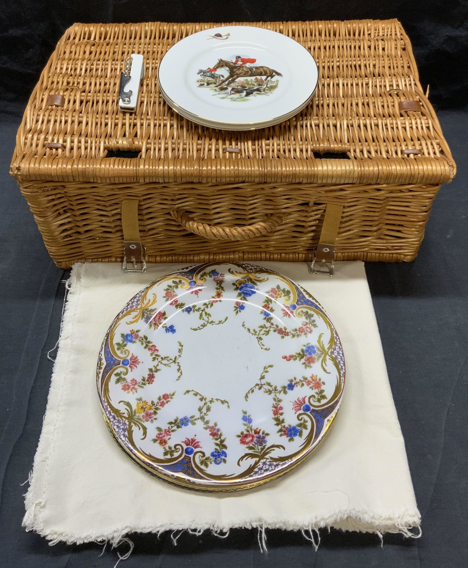 Lot 13 Picnic Basket, Plates, Tablecloth, More: Lot of 13 items. Wicker picnic basket with leather straps. Measures approx. 19 x 13 x 7 inches. 2 Thomas B Swain & Sin porcelain plates with horse and jockey motif. Measure approx. 7.75 inches diamete