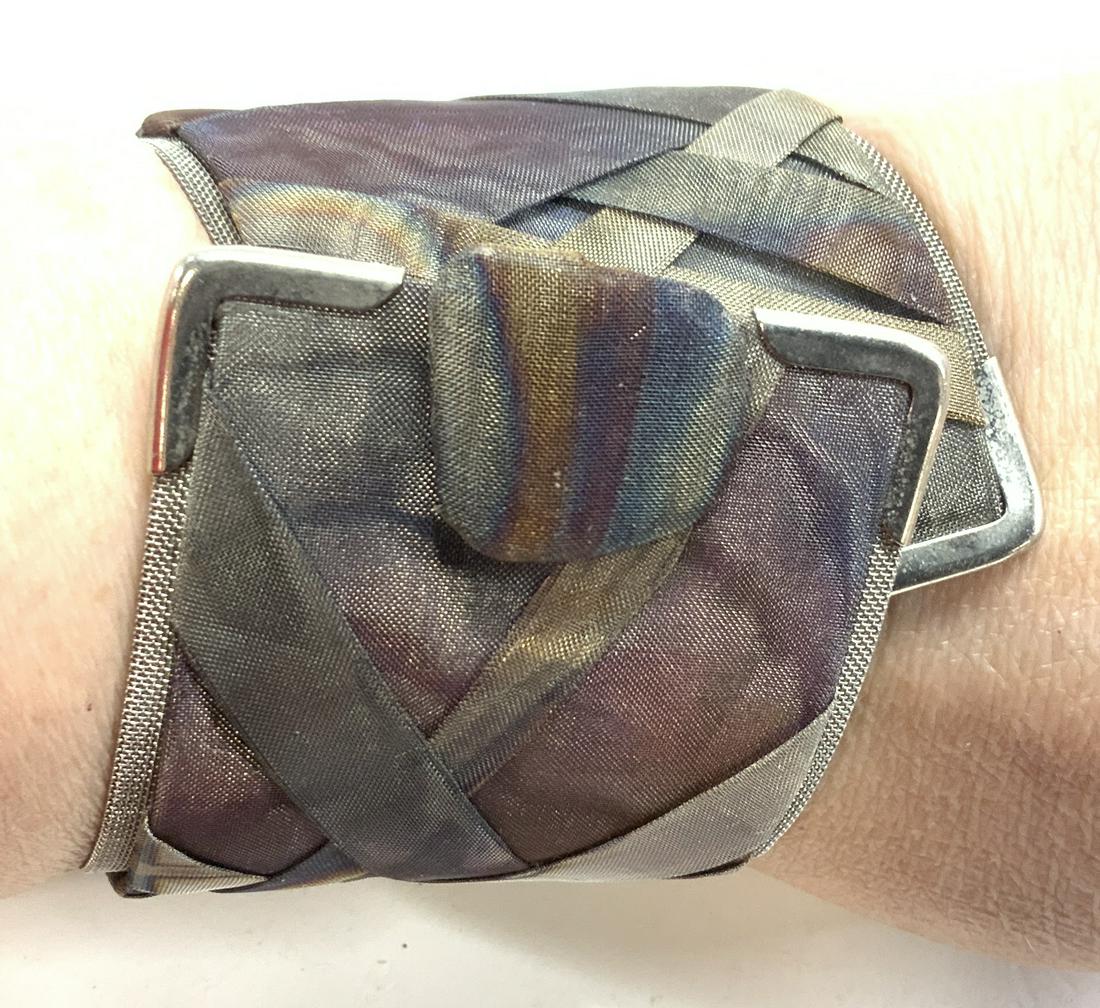 Artisanal Wrap Bracelet wit Mesh and Sheer Fabric: Wrap bracelet with snap closure, off set shape at ends, underside made with a metal mesh, exterior made with braided sheer fabric in silver and plumb colors. 2 in. Wide, 8.5 in. Long. Chunky jewelry, 