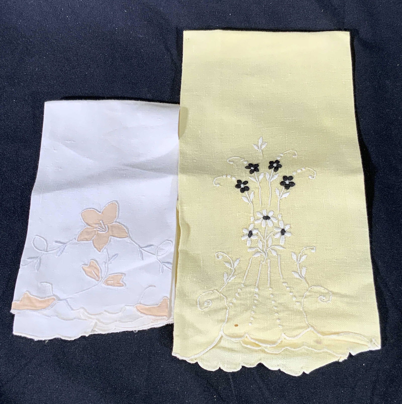 Lot 2 Vintage Embroidered Floral Tea Towels: Two vintage tea towels. Both pieces have scalloped edges. One is white toned with light apricot hued florals amongst white embroidery and pierced linen. Yellow hued tea towel features black and white