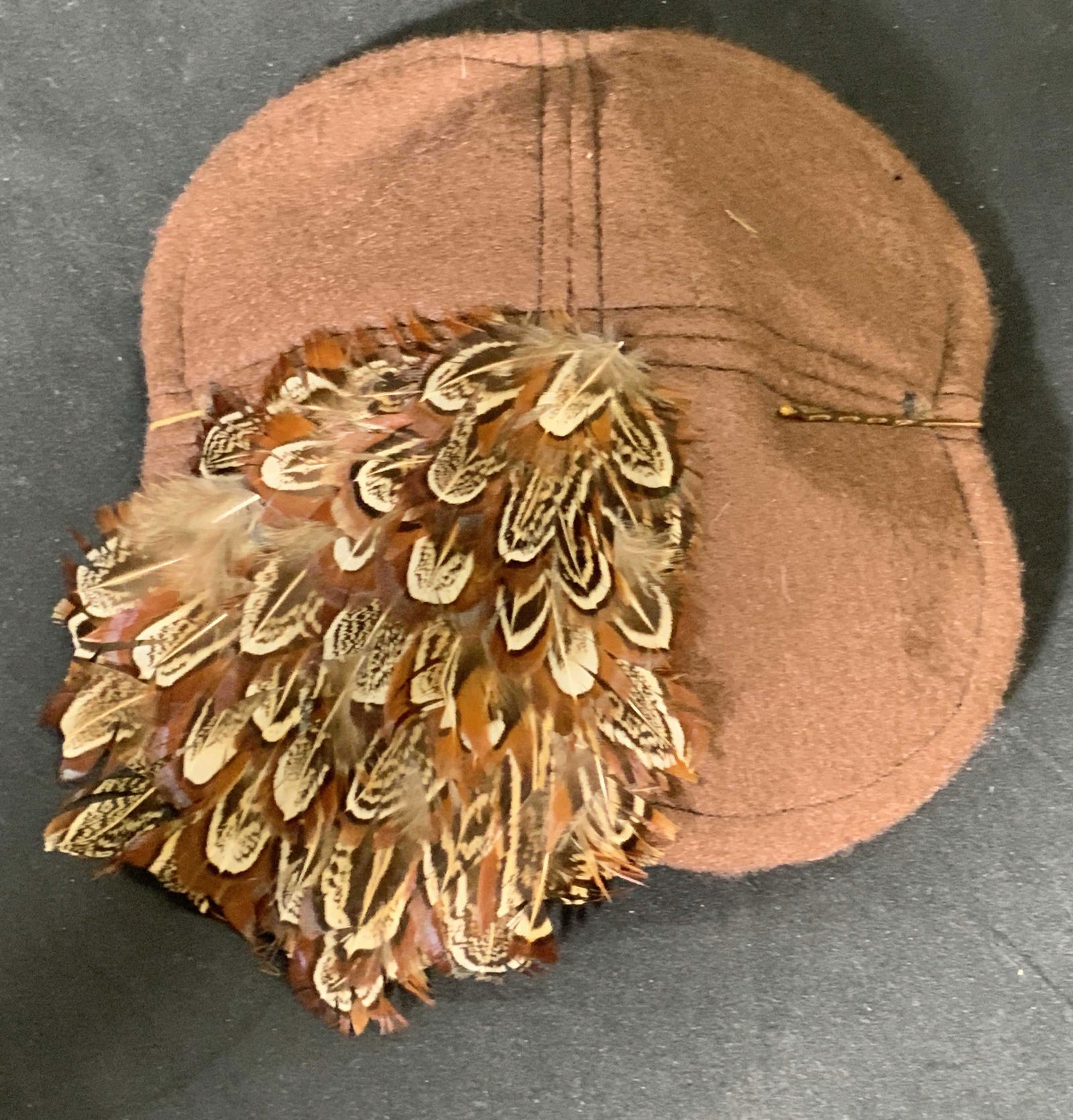 HUMBLEBUMBLEB Feathered Felt Cap: Brown felt cap featuring a feather detail to one side. Pins on either side of the cap. Measures approx 6.5L x 6.5W in. Tag to underside HUMBLEBUMBLEB.COM. Some wear consistent with use. Caps, hats, we