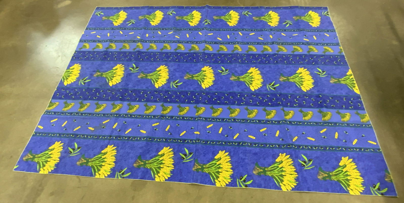 Wheat & Olive Harvest Motif Tablecloth: A tablecloth decorated with a wheat & olive pattern. Primarily blue, yellow, black, and green hues. Some discoloration present. Some wear consistent with use. Measures approx 77L x 59.5W in. No materi
