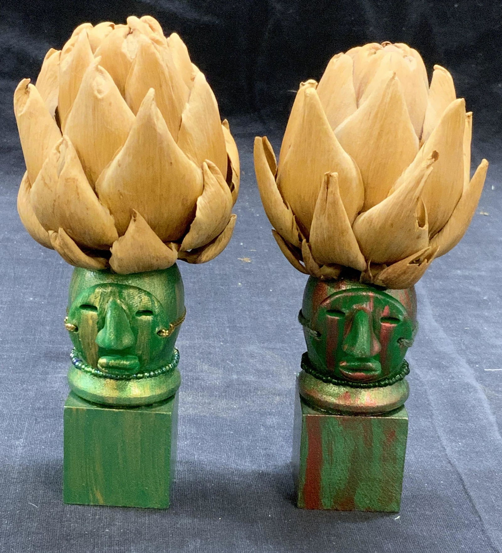 Pair Tribal Figural Artichoke Table Accessories: Pair of green and red hued tribal wood Figurals with dried artichoke decoration. Measure approx. 2.5 x 5.5 inches each. Some surface wear to ea h consistent with exposure. Tribal figural, artichoke, d