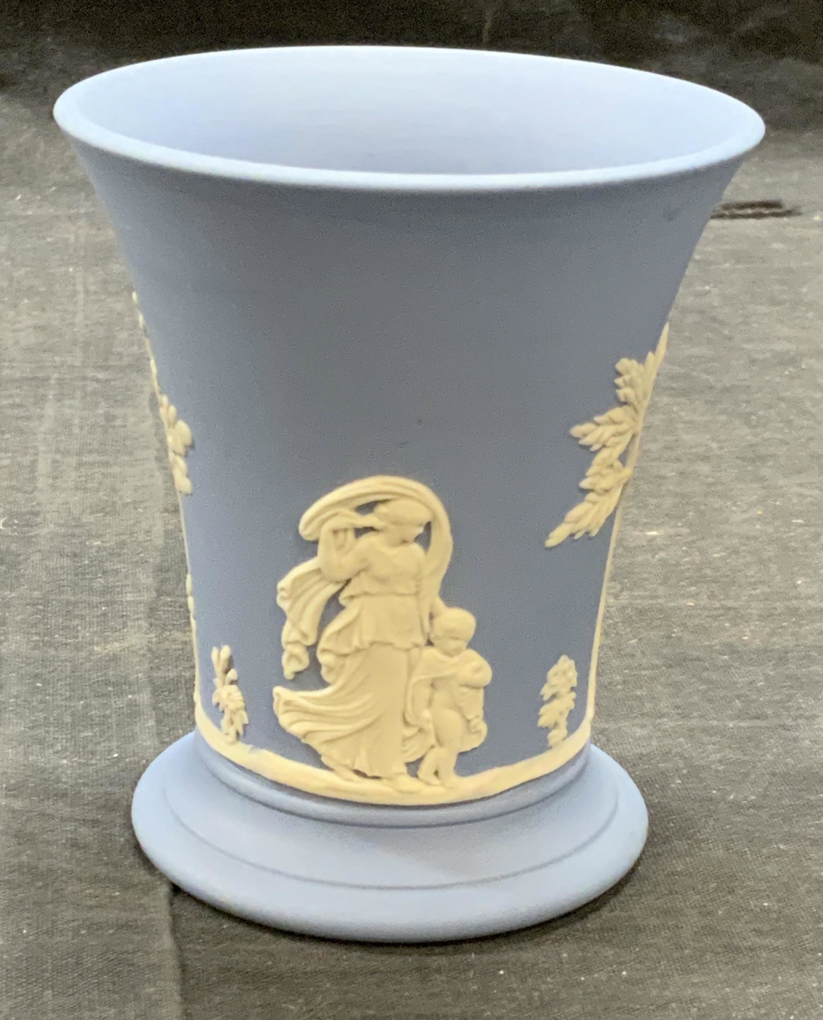 Wedgwood Blue Jasperware Grecian Figural Vase Eng: Wedgwood blue Jasperware vase with figural Grecian motif. Trademarked to underside. Made in England. Some surface wear consistent with exposure. Measures approx. 3 3/8 inches wide and 4 inches tall. W