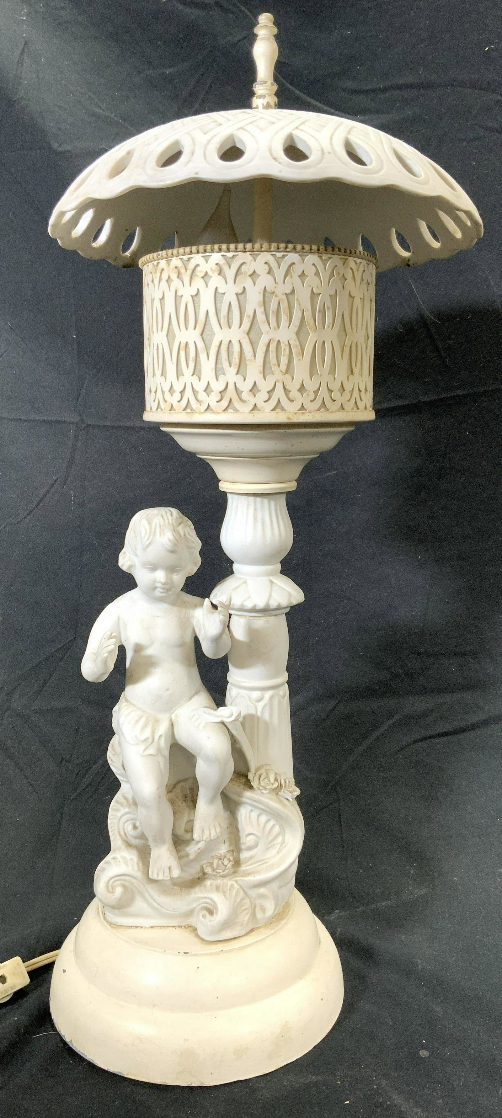 Vintage Porcelain & Metal Cherub Lamp: Lamp is vintage possibly antique. Piece is white toned, porcelain & metal. Piece has umbrella style shade with pierced detail. Base has seated cherub figure. Lamp measures approx 20 inches tall 9 inch