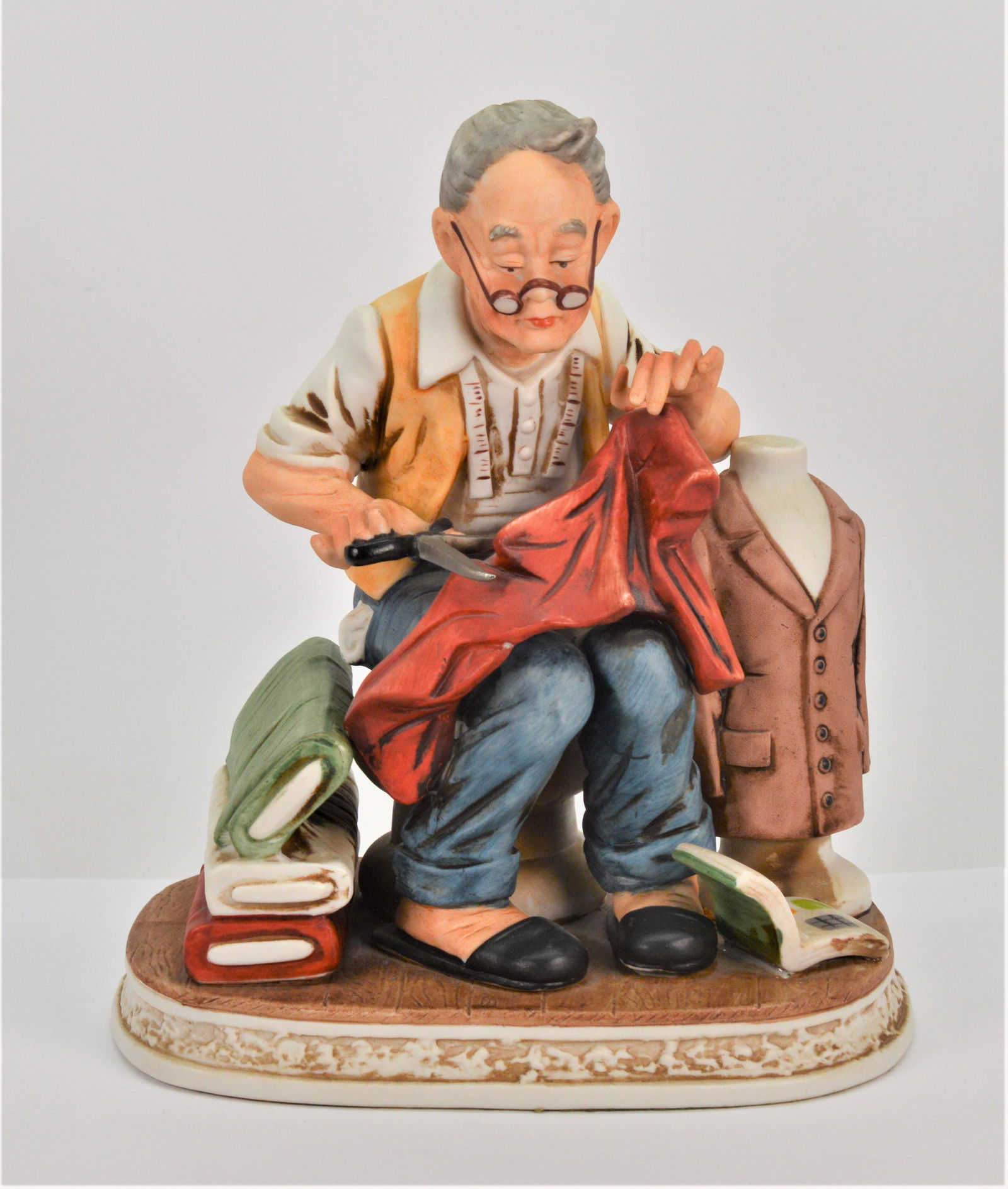 Bisque Porcelain Statue of a Tailor: This statue depicts an older gentlemen tailor hard at work with his scissors and pattern book. This stands approximately 8 high 6.5 wide and 4.75 deep. No visible nicks or chips.-nb