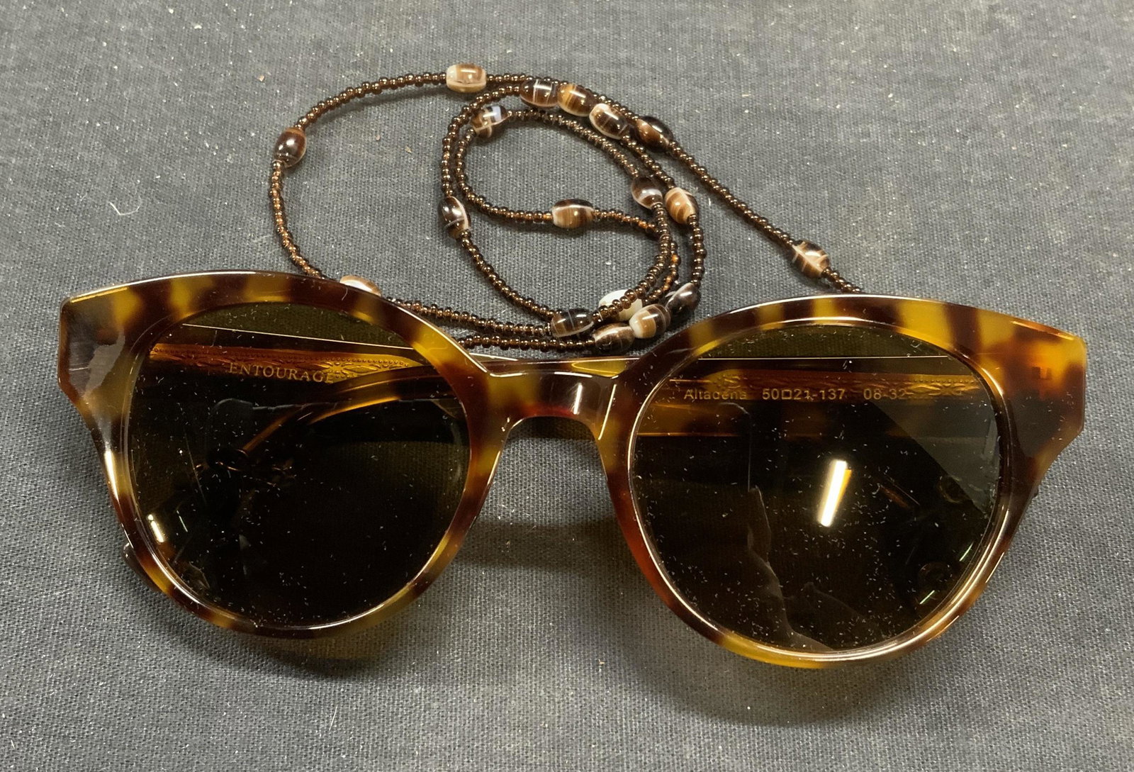 Entourage of 7 Brown Sunglasses with Chain: Entourage of 7 brown tortoise pattern acrylic frame sunglasses with beaded glasses chain. Designer label to arm. Measure approx. 5 x 5.5 x 2 inches. Includes sunglasses case. Some surface wear consist