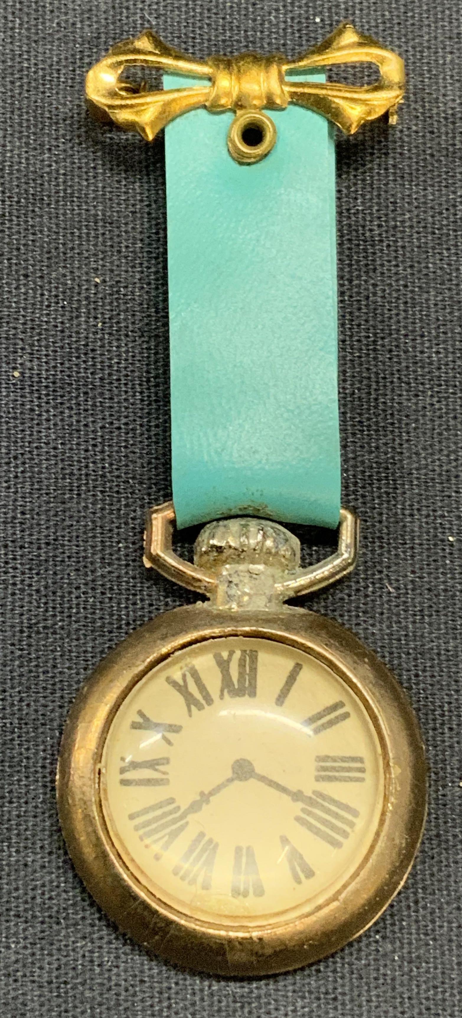 Faux Gold Tone Pocket Watch Brooch: Gold tone faux pocket watch brooch with blue strap and bow decoration. Hinge pin to underside. Surface wear consistent with exposure. Measures approx. 2.75 x 1 inches. Estate jewelry, costume jewelry,