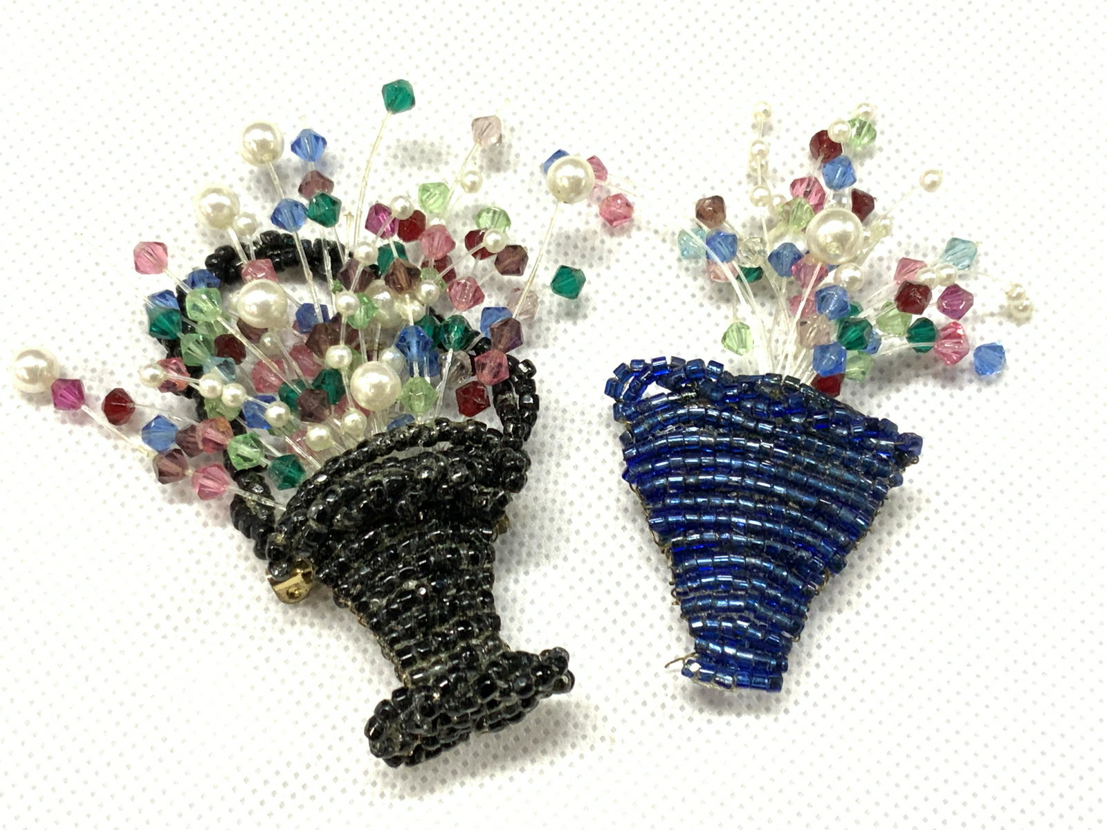 2 Arts & Crafts Basket Of Flowers Brooches: 2 assorted arts and crafts brooches, each made with woven seed beads with bouquets of stringed beads to resemble flowers. 1 depicts a black basket and the other is a blue vase. Largest is approximatel