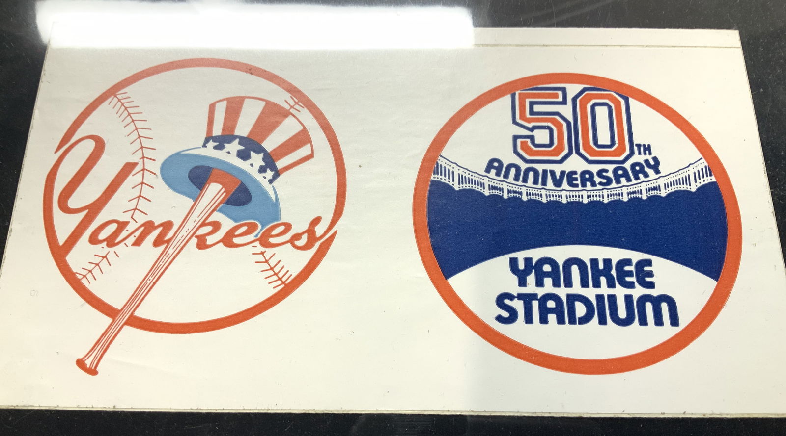 50th Anniversary Yankee Stadium Bumper Sticker: 8 x 4 1/8 in. Bumper sticker of 2 Yankees Logos. One Yankees logo with Americana Hat on Baseball and 50th Anniversary Yankee Stadium. Stored in heavy plastic folder. Property of 5th Avenue Manhattan E