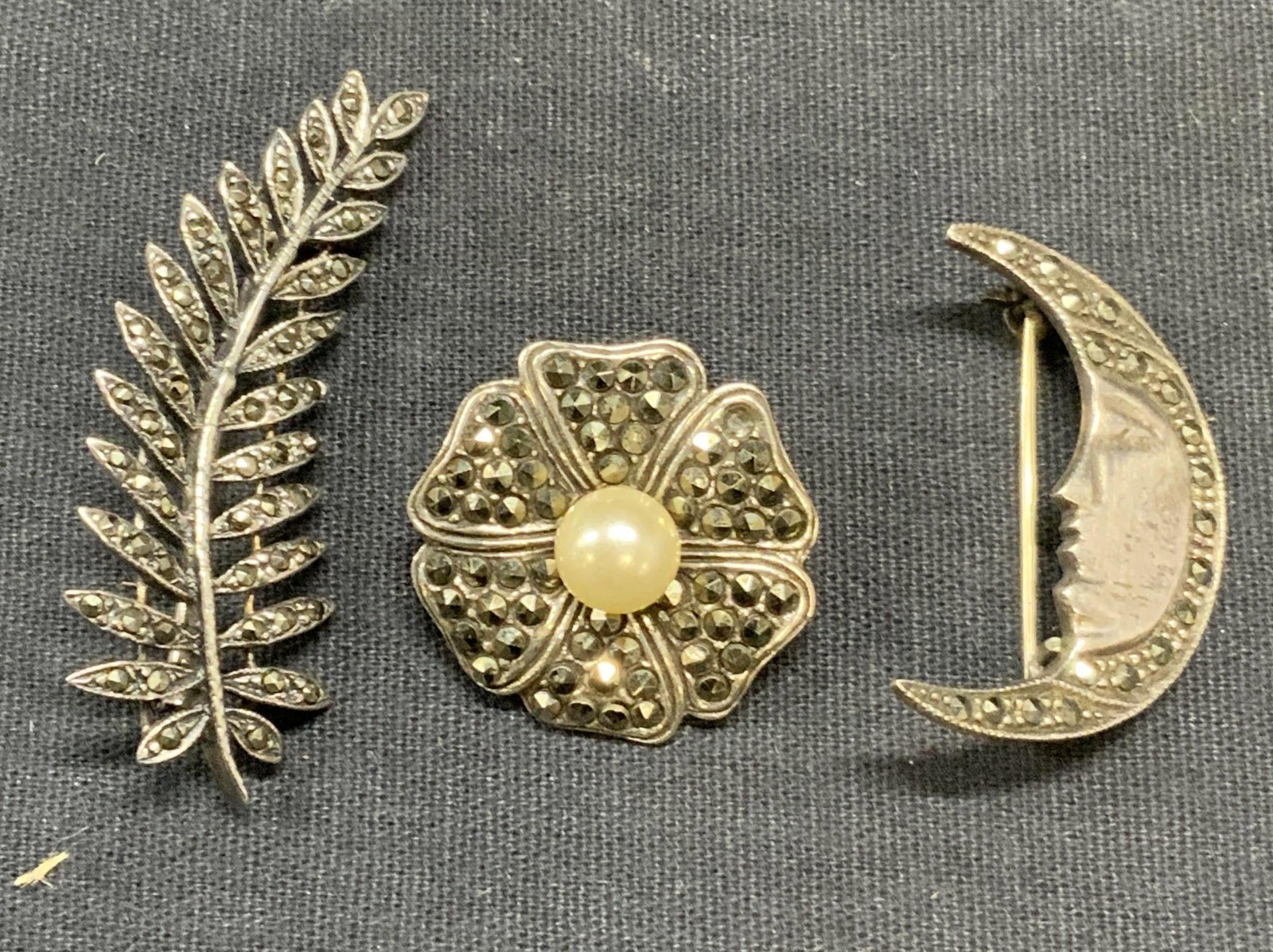Lot 3 Sterling Silver & Marcasite Brooches: Lot of 3 sterling silver and marcasite brooches. Flower brooch with faux pearl decoration, marked 925 and hallmarked to underside. Measures approx. 1 inch diameter. Mood figural brooch. Measures appro
